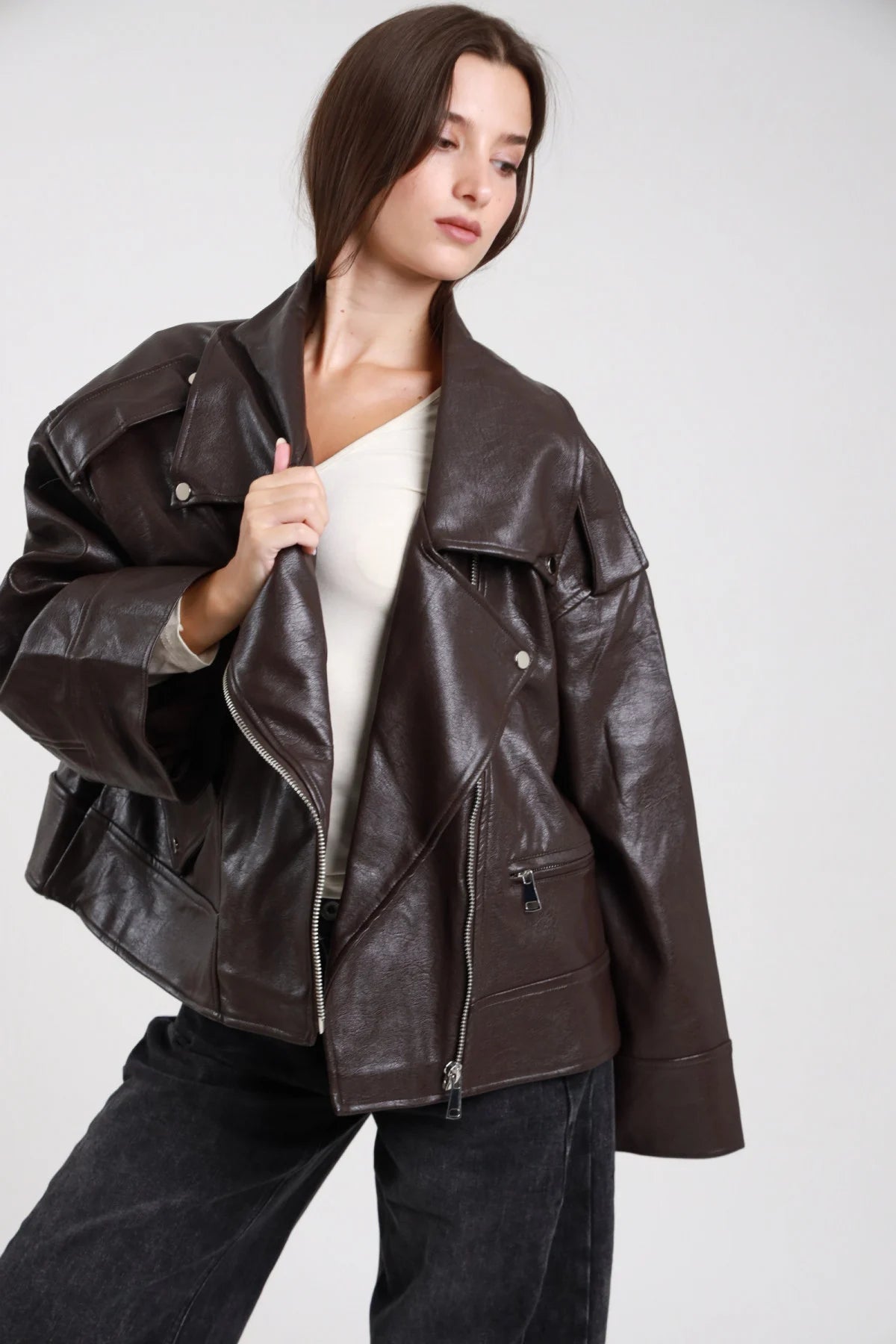 Chiodo over ecopelle marrone cioccolato scuro donna oversize colletto revers ampio zip asimmetrica argento texture crinkle - vista frontale tre quarti mano al colletto aperto con jeans neri