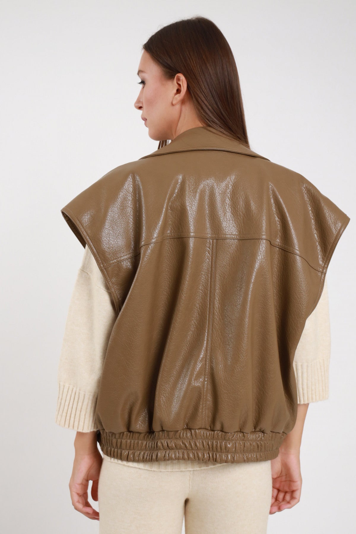 Chiodo Ecopelle Smanicato taupe kaki verde verniciata crinkled revers biker drop shoulder vita elastica arricciata retro intera schiena - modella retro intera sguardo laterale capelli castani lisci sfondo bianco maglione beige pantalone beige