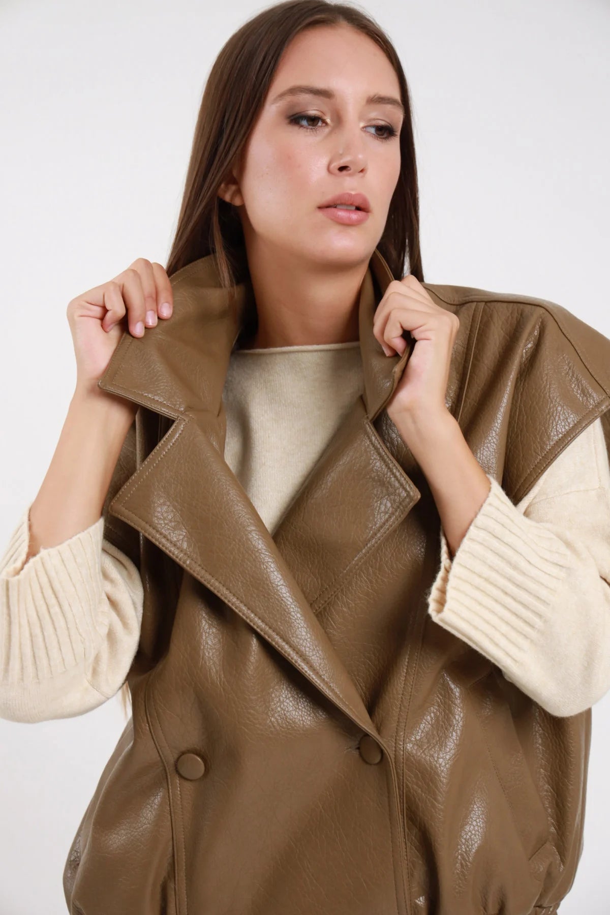 Chiodo Ecopelle Smanicato taupe kaki verde verniciata crinkled revers biker ampio doppiopetto drop shoulder vita elastica busto frontale 3/4 mani revers aperto - modella busto frontale 3/4 mani revers sguardo alto capelli castani lisci sfondo bianco maglione beige