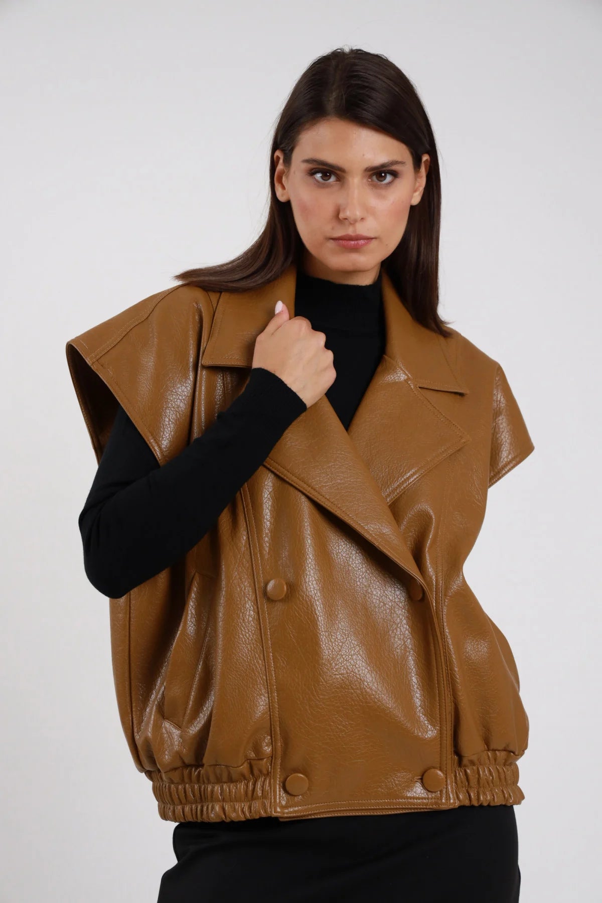 Chiodo Ecopelle Smanicato cuoio cammello verniciata crinkled revers biker ampio doppiopetto drop shoulder vita elastica busto frontale 3/4 mano revers sguardo intenso - modella busto frontale 3/4 mano revers sguardo frontale capelli castani lisci sfondo bianco dolcevita nero