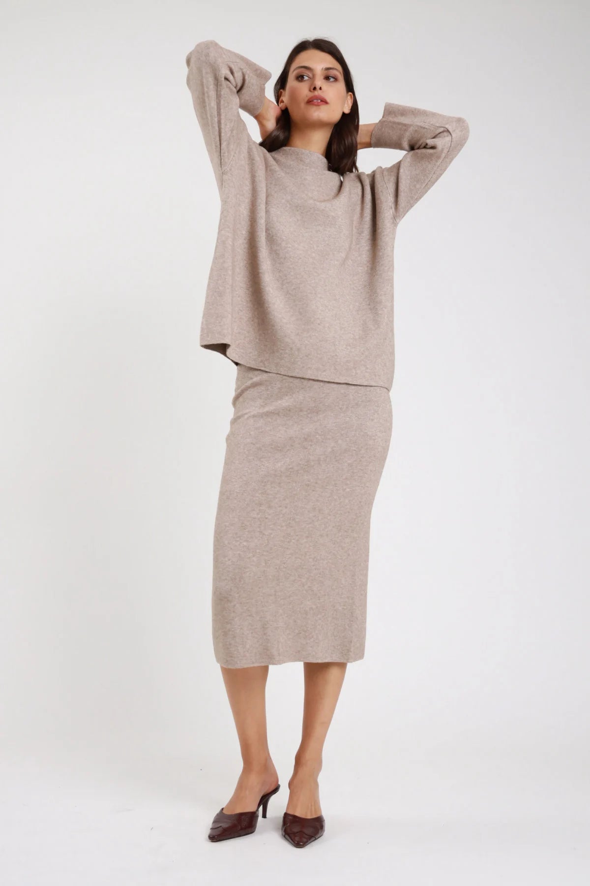 Completo coordinato gonna midi pencil coste rib maglia oversize batwing mock neck beige sabbia melange frontale intera braccia alzate testa - modella frontale intera braccia alzate testa sguardo frontale capelli castani lisci raccolti sfondo bianco mules tacco beige