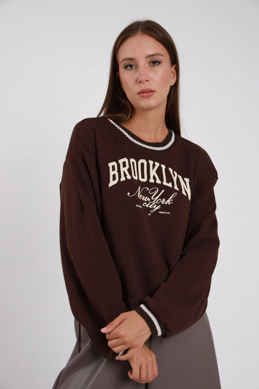 Felpa Brooklyn marrone cioccolato stampa varsity college avorio collo rotondo bordo contrasto avorio bianco polsini contrasto oversize drop shoulder busto frontale 3/4 mani giunte sguardo camera - modella busto frontale 3/4 mani giunte sguardo camera capelli castani lisci sfondo bianco gonna grigio talpa