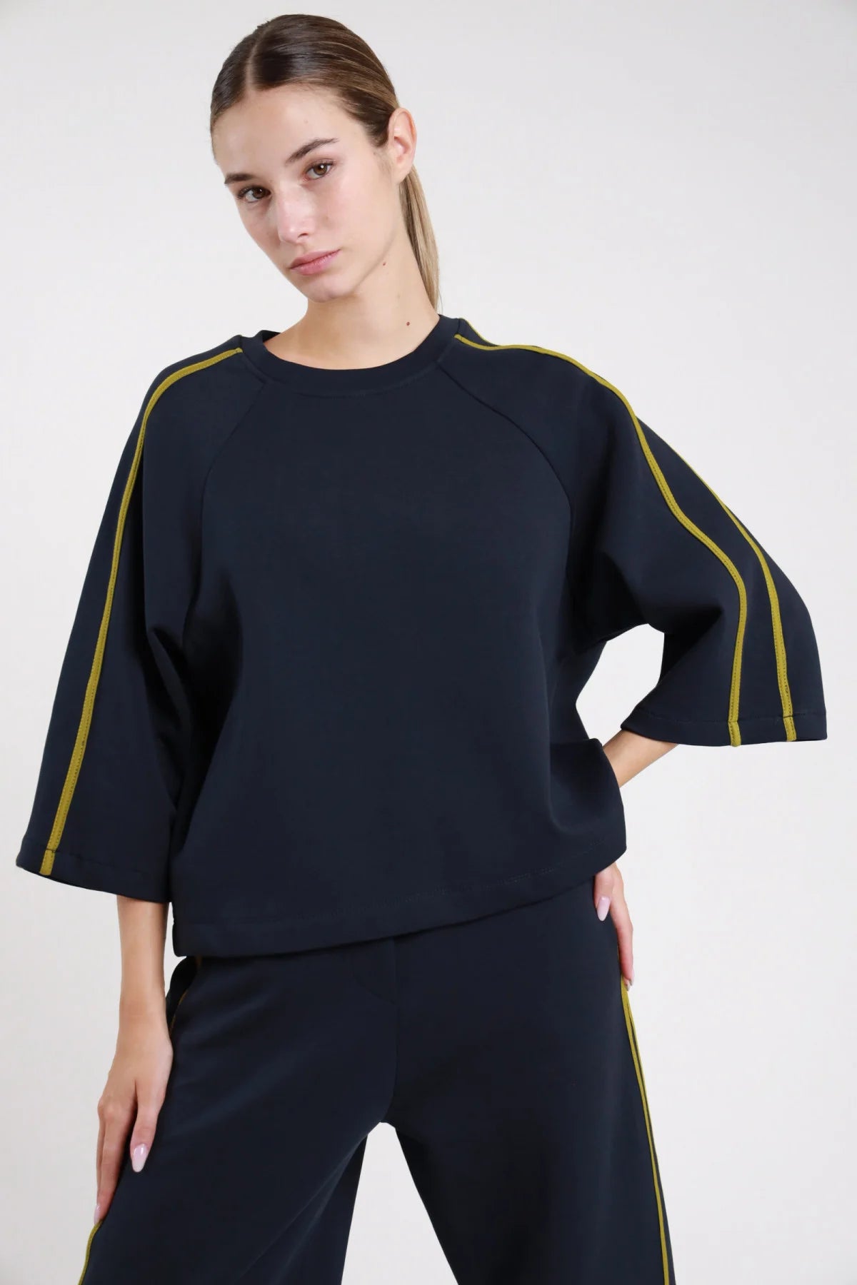 Felpa bande nera frontale busto mano fianco bande verde khaki verticali spalla manica bat sleeve corpo boxy maxi - modella frontale busto mano fianco sguardo frontale pantaloni neri coordinato capelli castani raccolti