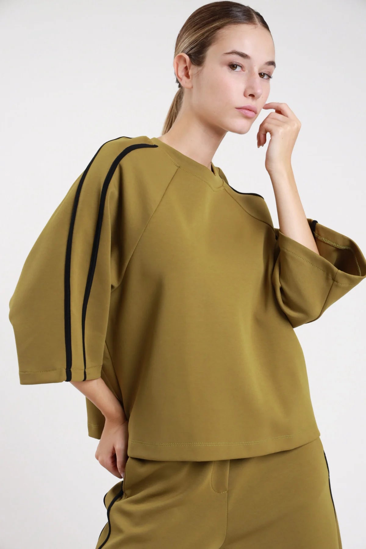 Felpa bande verde khaki oliva frontale busto tre quarti bande nere verticali spalla manica bat sleeve oversize - modella frontale busto tre quarti mano mento sguardo laterale pantaloni khaki coordinato capelli castani lisci raccolti