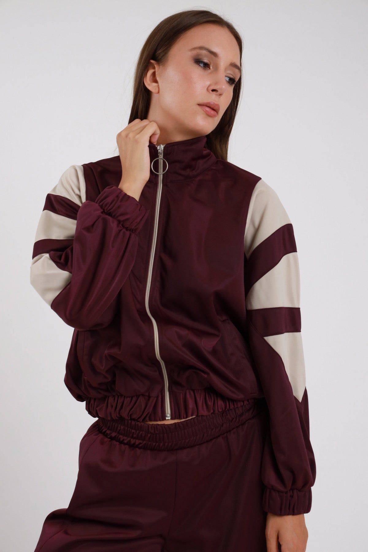 Giacca Zip Bande Diagonali bordeaux prugna raso satin collo alto rialzato bande diagonali panna larghe obliquo maniche zip anello argento elastico fondo bomber crop - modella busto 3/4 laterale posa mano mento sguardo laterale capelli castani lisci sfondo bianco pantalone palazzo bordeaux coordinato