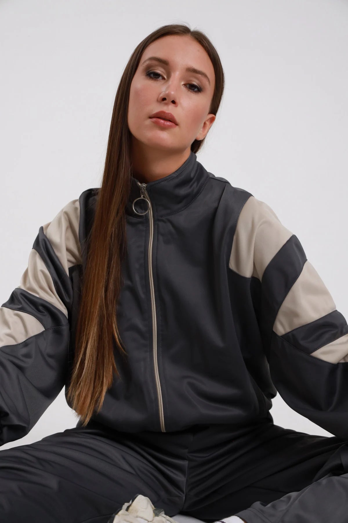 Giacca Zip Bande Diagonali antracite grigio scuro raso satin collo alto rialzato bande diagonali panna larghe obliquo maniche zip anello argento elastico fondo bomber crop polsini elastici - modella busto 3/4 frontale seduta posa sguardo frontale intenso make-up bold capelli castani lisci sfondo bianco pantaloni palazzo antracite coordinati sneakers bianche