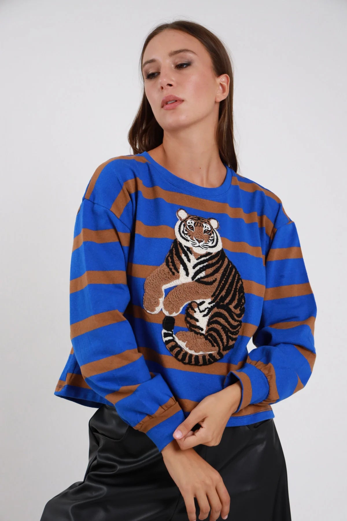 Felpa Girocollo Righe Larghe Patch Tigre blu elettrico cobalto marrone cammello 77% cotone maxi patch tigre ricamata realistica multicolor centro petto manica ampia polsino elastico oversize boxy - modella busto 3/4 frontale seduta posa mani grembo sguardo basso soft capelli castani lisci sfondo bianco gonna ecopelle nera lucida