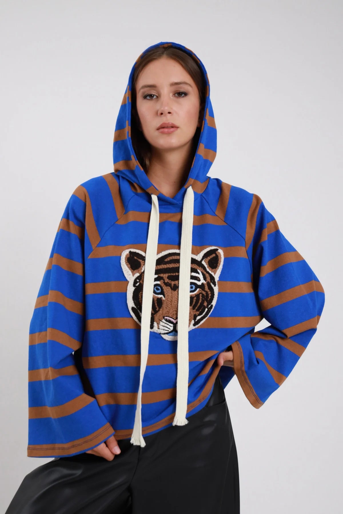 Hoodie Righe Larghe Patch Testa Tigre blu elettrico cobalto marrone cammello 77% cotone cappuccio indossato cordini panna lunghi maxi patch testa tigre ricamata multicolor petto manica batwing ampia silhouette maxi boxy crop - modella busto frontale posa mano fianco sguardo frontale intenso capelli castani lisci sfondo bianco pantaloni ecopelle nera