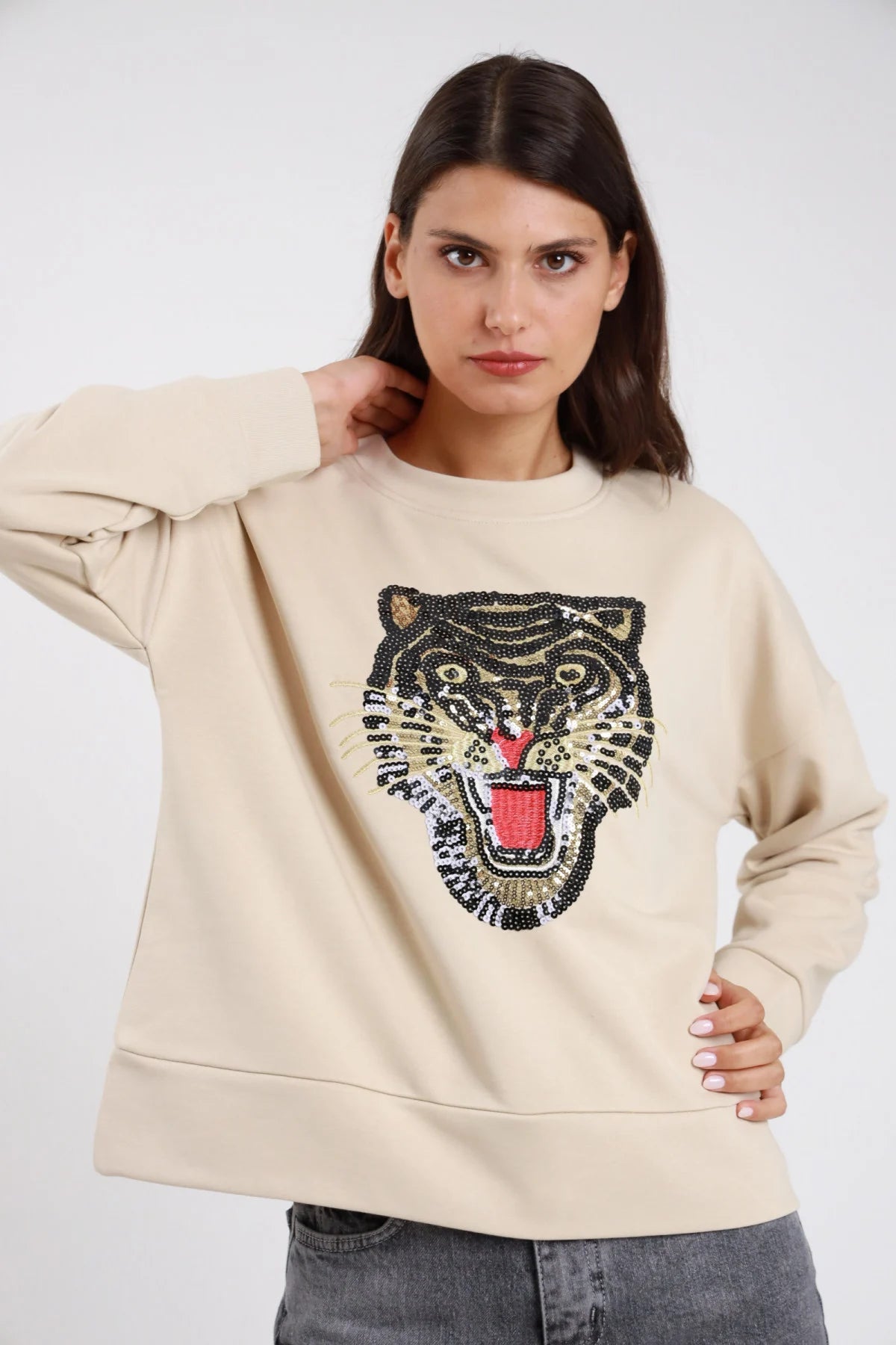 Felpa oversize cotone felpato beige sabbia crema maxi patch ricamo paillettes tigre ruggente oro nero rosso girocollo tondo frontale busto mano fianco - modella frontale busto mano fianco sguardo frontale capelli castani scuri lisci corti sfondo bianco jeans grigio