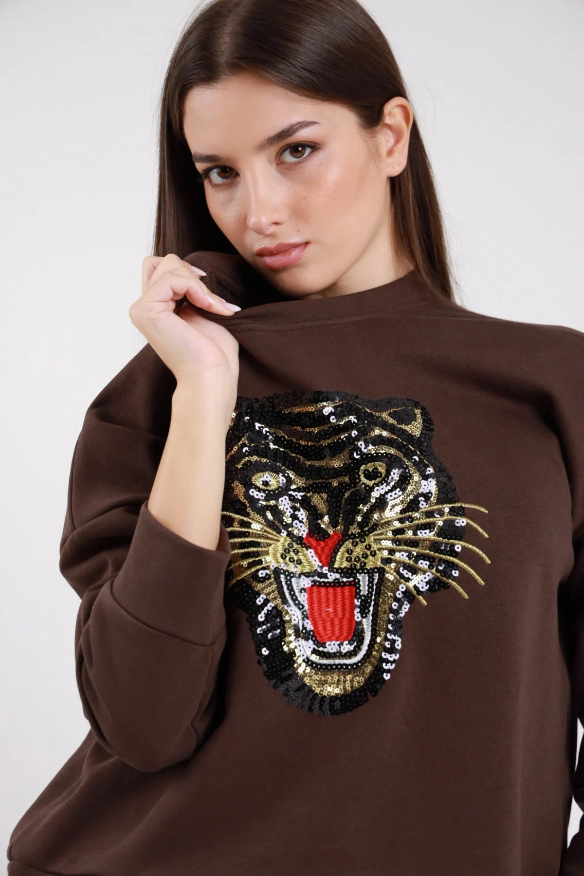 Felpa oversize cotone felpato marrone cioccolato maxi patch ricamo paillettes tigre ruggente oro nero rosso girocollo tondo frontale busto tigre close-up mano spalla - modella frontale busto mano spalla sguardo frontale capelli castani scuri lisci lunghi sfondo bianco pantaloni check beige