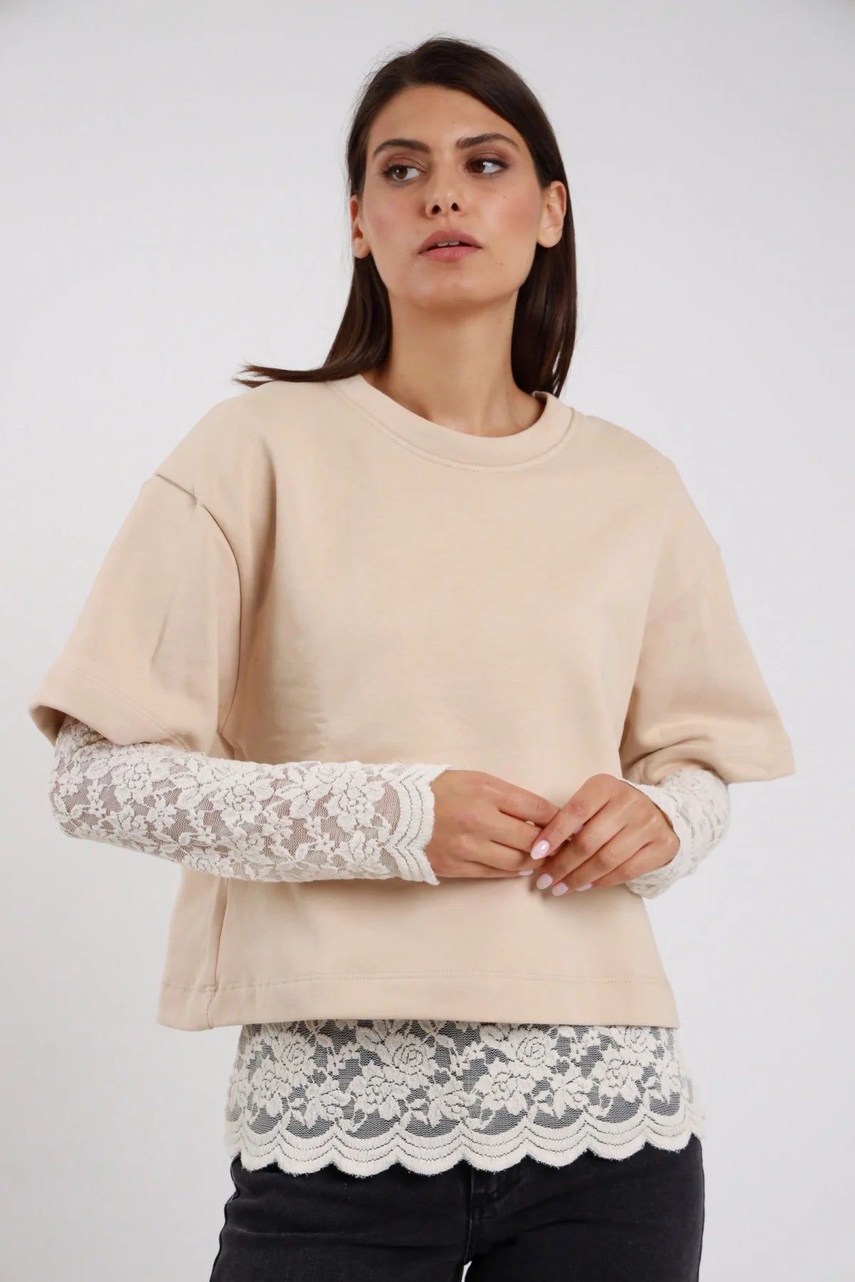 Felpa Girocollo Oversize Inserto Pizzo Floreale beige sabbia 100% cotone girocollo costina manica 3/4 inserto pizzo bianco panna polso avambraccio orlo pizzo smerlato bianco silhouette boxy - modella busto frontale mani intrecciate sguardo laterale alto capelli castani lisci sfondo bianco jeans neri
