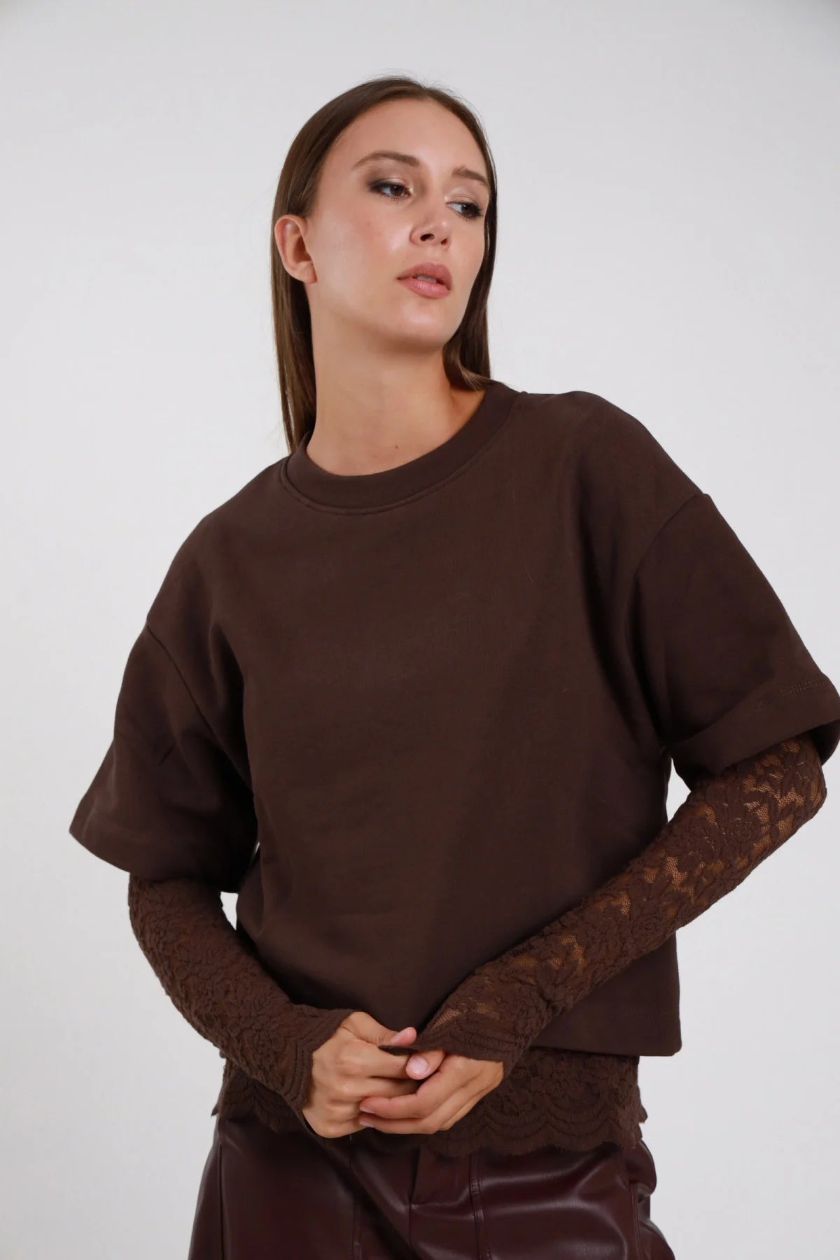 Felpa Girocollo Oversize Inserto Pizzo Floreale marrone cioccolato 100% cotone girocollo costina manica 3/4 inserto pizzo marrone tono su tono polso avambraccio silhouette boxy crop - modella busto frontale mani intrecciate sguardo frontale capelli castani lisci sfondo bianco pantaloni ecopelle marrone coordinati