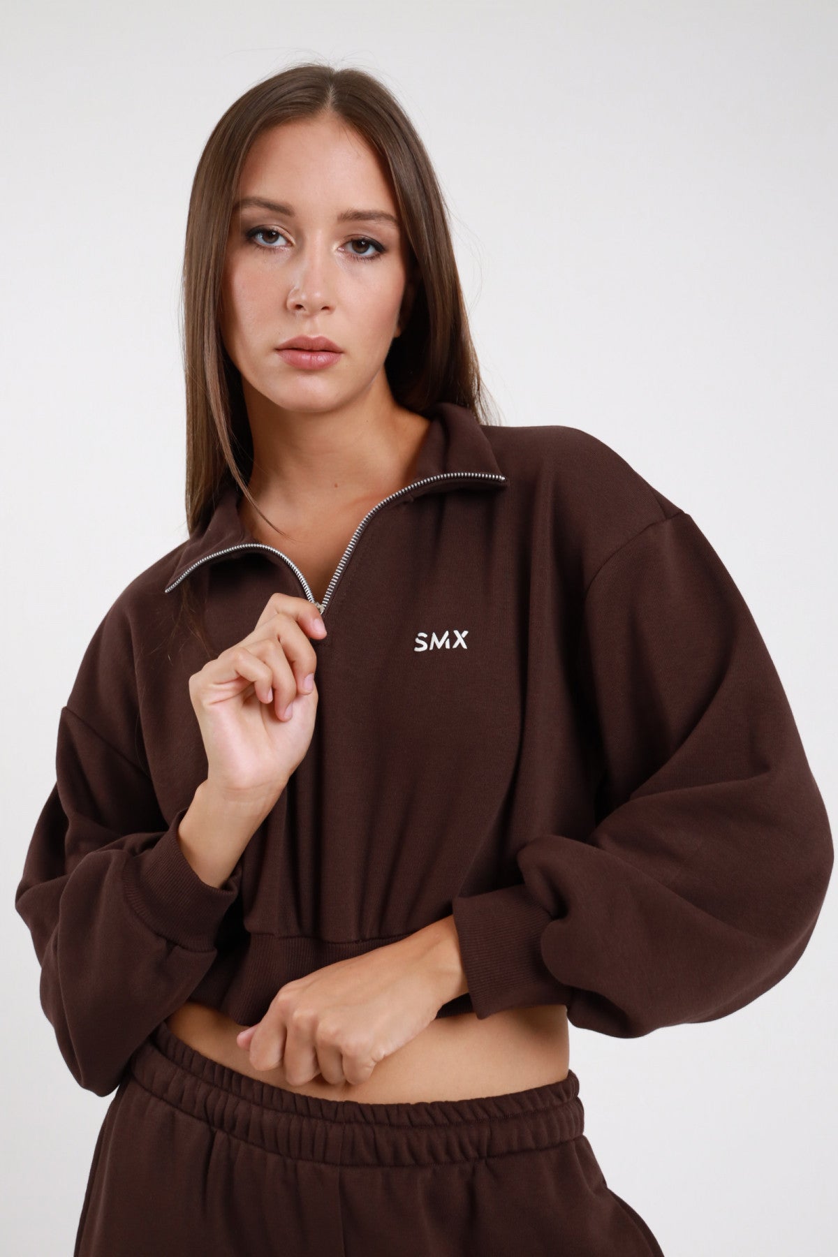 Felpa Crop Half-Zip DAYTHRU marrone cioccolato scuro colletto alto lupetto zip 1/2 dorata logo SMX ricamato bianco petto manica oversize balloon orlo elastico arricciato crop busto frontale 3/4 mano zip sguardo frontale - modella busto frontale 3/4 mano zip sguardo frontale capelli castani lisci sfondo bianco pantalone tuta jogger marrone coordinato