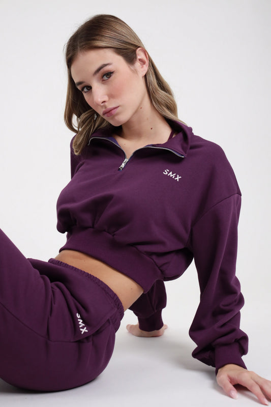 Felpa Crop Half-Zip SMX Limited Edition viola melanzana 65% cotone colletto ribaltato polo zip 1/2 logo SMX bianco petto manica balloon polsino costina bordo costina crop seduta 3/4 laterale sguardo intenso - modella seduta accovacciata 3/4 laterale sguardo intenso capelli castani lisci sfondo bianco pantalone jogger viola SMX coordinato abbinato