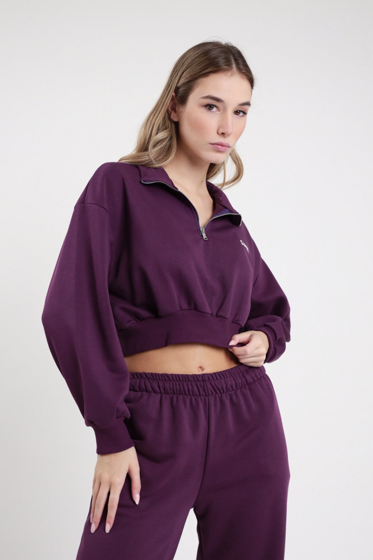 Felpa Crop Half-Zip SMX Limited Edition viola melanzana 65% cotone colletto ribaltato zip 1/2 logo SMX petto bordo costina crop frontale busto mano bordo fondo - modella busto frontale mano bordo fondo sguardo laterale capelli castani sfondo bianco pantalone jogger viola SMX coordinato abbinato