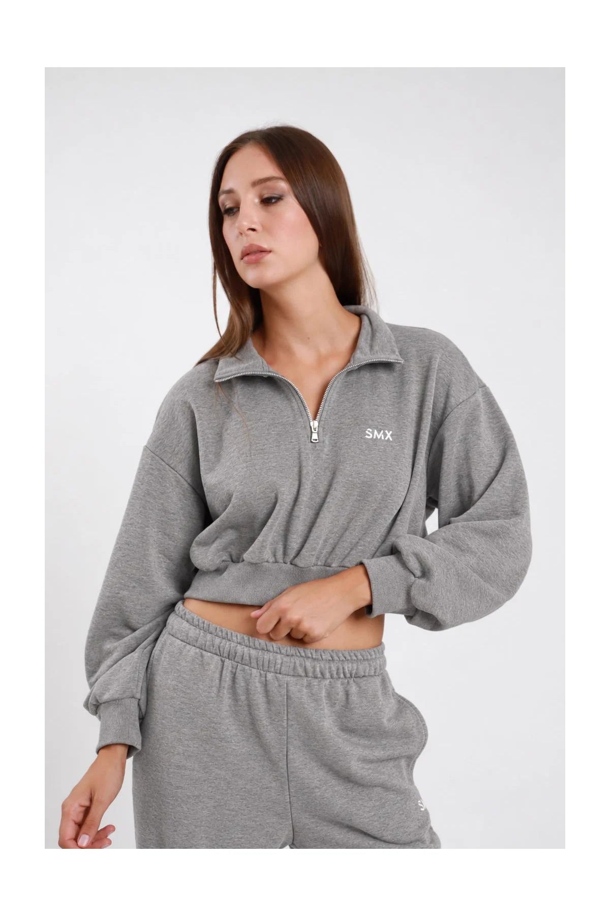 Felpa Crop Half-Zip DAYTHRU grigio melange chiaro colletto alto lupetto zip 1/2 argento logo SMX ricamato bianco petto manica oversize balloon orlo elastico arricciato crop busto frontale 3/4 mano zip sguardo basso - modella busto frontale 3/4 mano zip sguardo basso capelli castani lisci sfondo bianco pantalone tuta jogger grigio coordinato