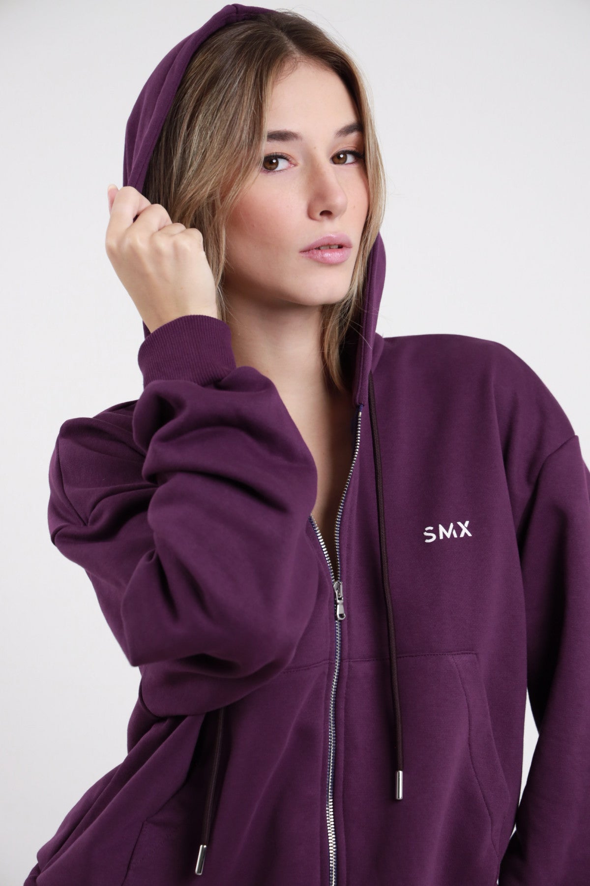 Felpa Zip SMX Limited Edition viola melanzana 65% cotone hoodie cappuccio coulisse zip frontale logo SMX bianco petto sinistro ricamato manica oversize balloon busto 3/4 mano cappuccio - modella busto 3/4 mano cappuccio sguardo frontale capelli castani lisci sfondo bianco pantalone wide leg viola coordinato abbinato