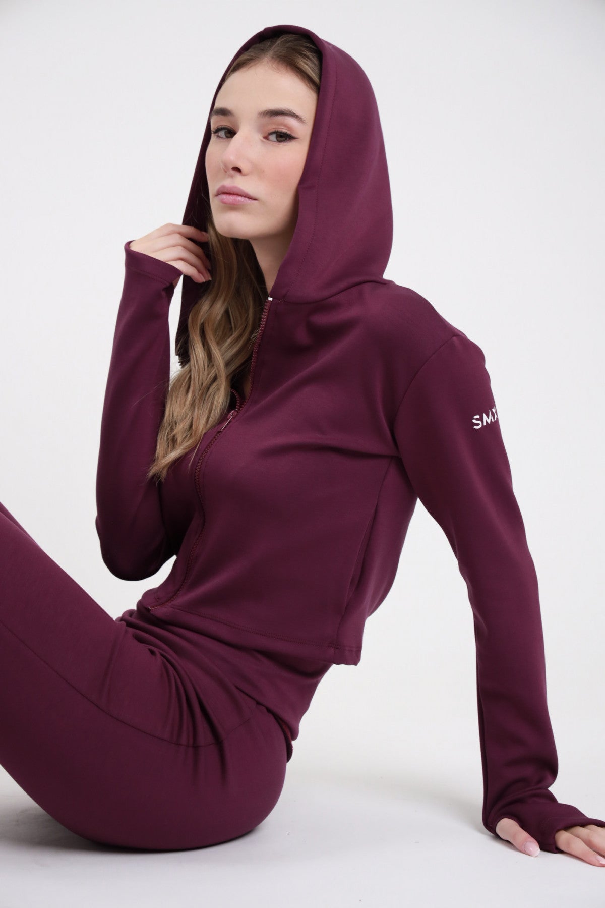 Felpa Crop Zip SMX Limited Edition bordeaux prugna 50% poliestere viscosa hoodie cappuccio coulisse zip frontale logo SMX bianco braccio thumbhole polsino crop fitted seduta 3/4 laterale mano cappuccio - modella seduta 3/4 laterale mano cappuccio sguardo frontale capelli mossi castani sfondo bianco pantaloni flare SMX bordeaux coordinati abbinati