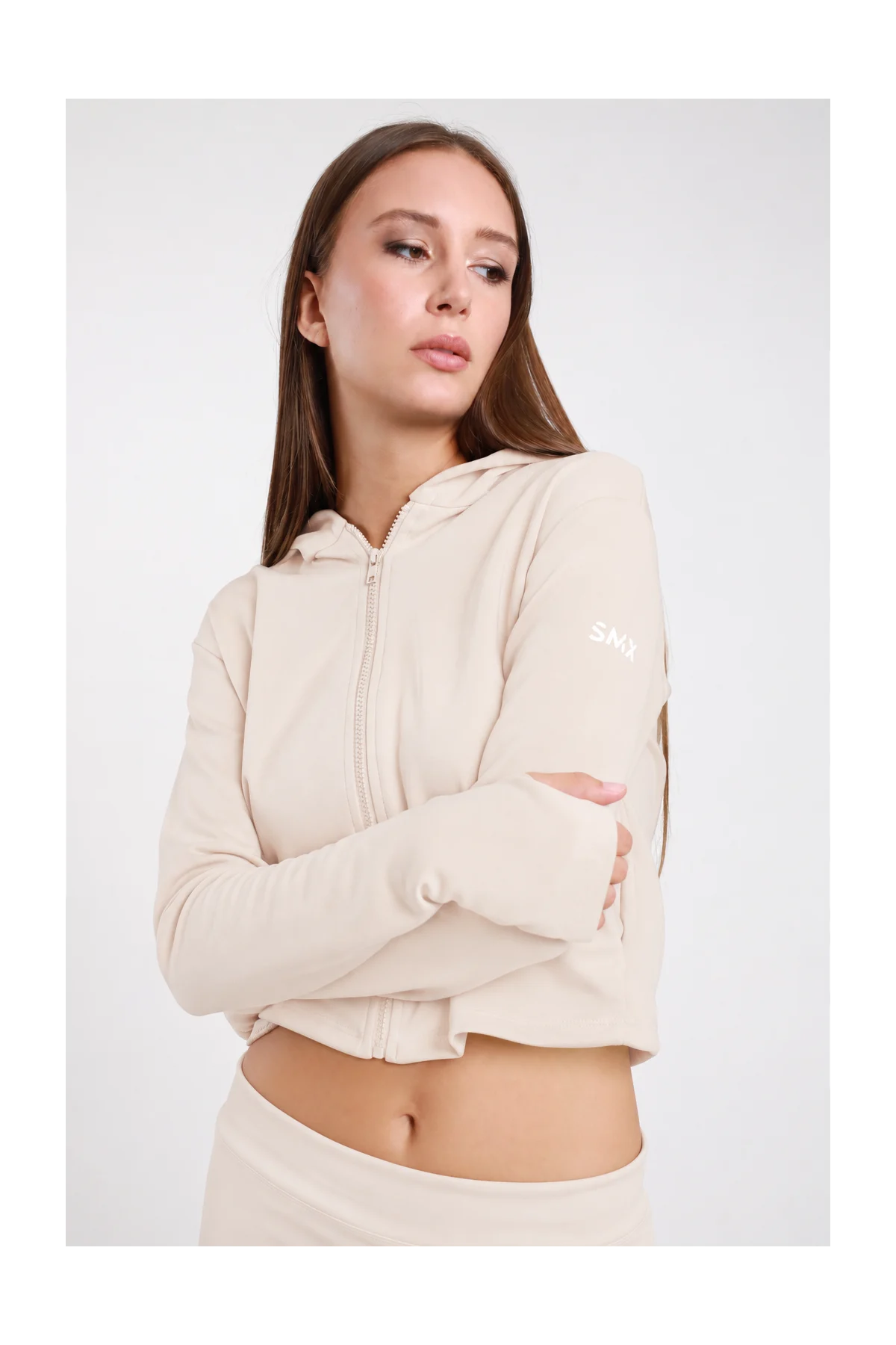Felpa Crop Zip DAYTHRU sabbia beige chiaro cappuccio zip frontale logo SMX bianco manica sinistra orlo crop polsino foro pollice crop fit busto frontale 3/4 braccia conserte sguardo frontale - modella busto frontale 3/4 braccia conserte sguardo frontale capelli castani lisci sfondo bianco coordinato sabbia beige