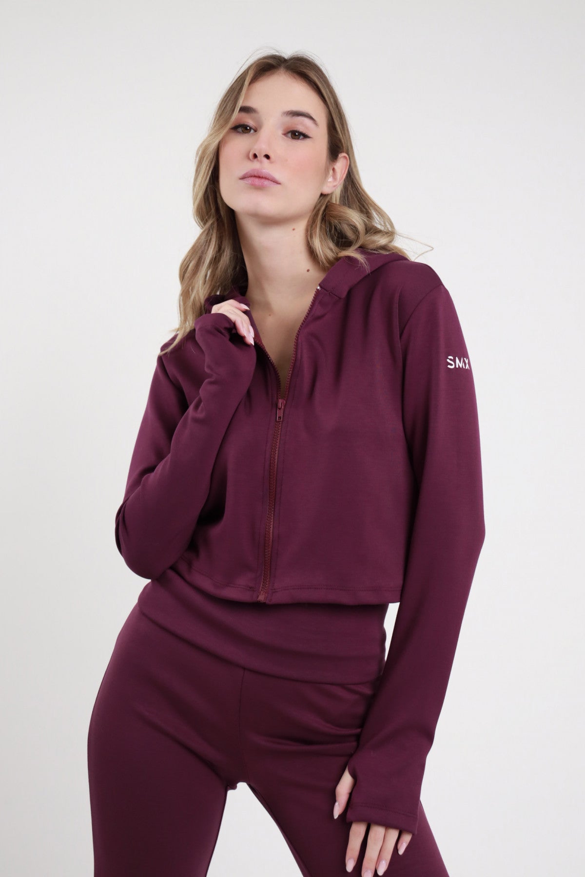 Felpa Crop Zip SMX Limited Edition bordeaux prugna cappuccio coulisse zip frontale aperta logo SMX braccio thumbhole crop fitted frontale busto mano cappuccio - modella busto frontale mano cappuccio sguardo frontale zip aperta capelli biondi mossi sfondo bianco pantaloni flare SMX bordeaux coordinati abbinati
