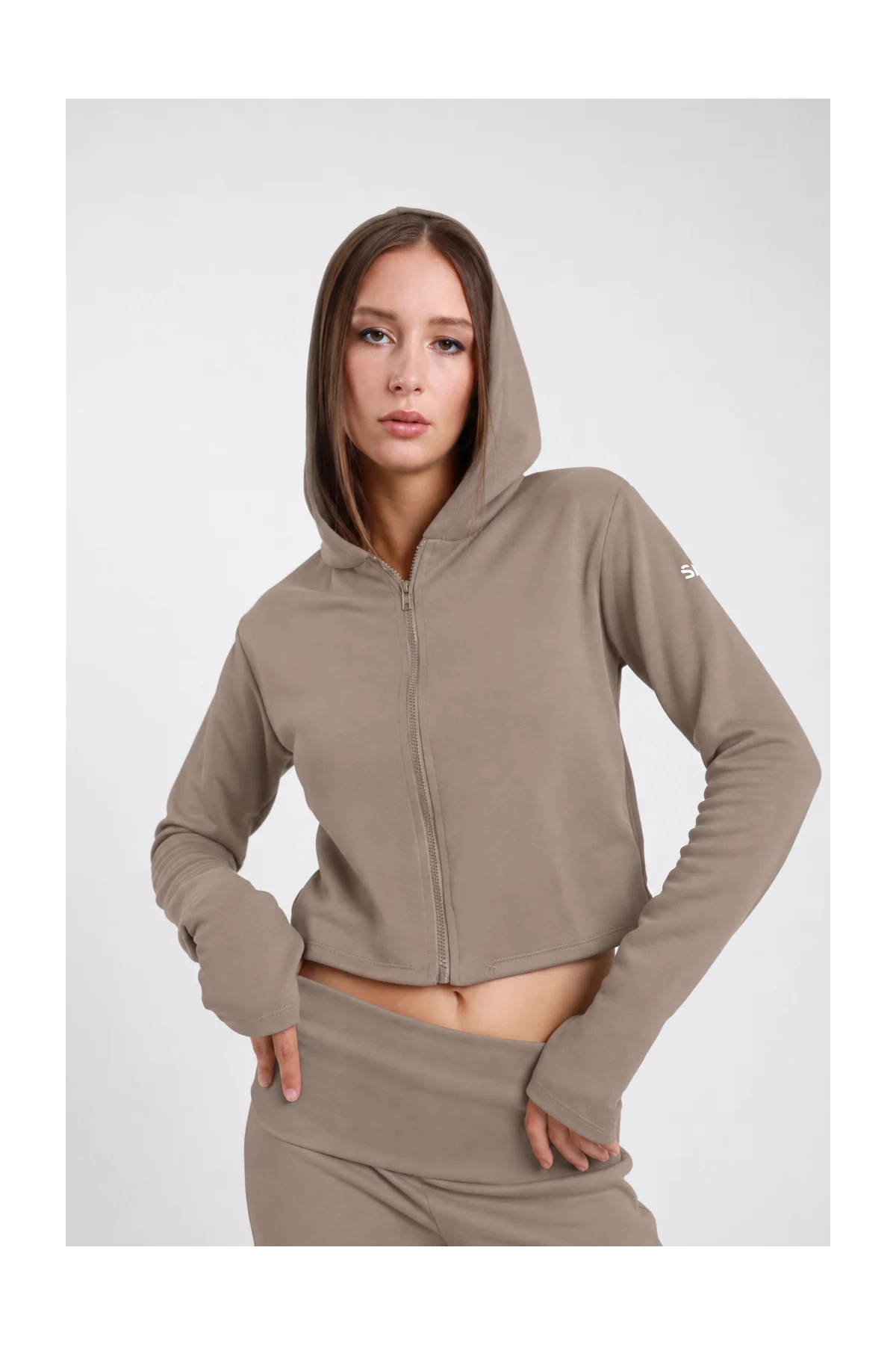 Felpa Crop Zip DAYTHRU grigio taupe tortora cappuccio zip frontale logo SMX bianco manica orlo crop polsino foro pollice crop fit busto frontale 3/4 mani fianchi sguardo frontale - modella busto frontale 3/4 mani fianchi sguardo frontale capelli castani lisci sfondo bianco coordinato grigio taupe pantaloni wide-leg