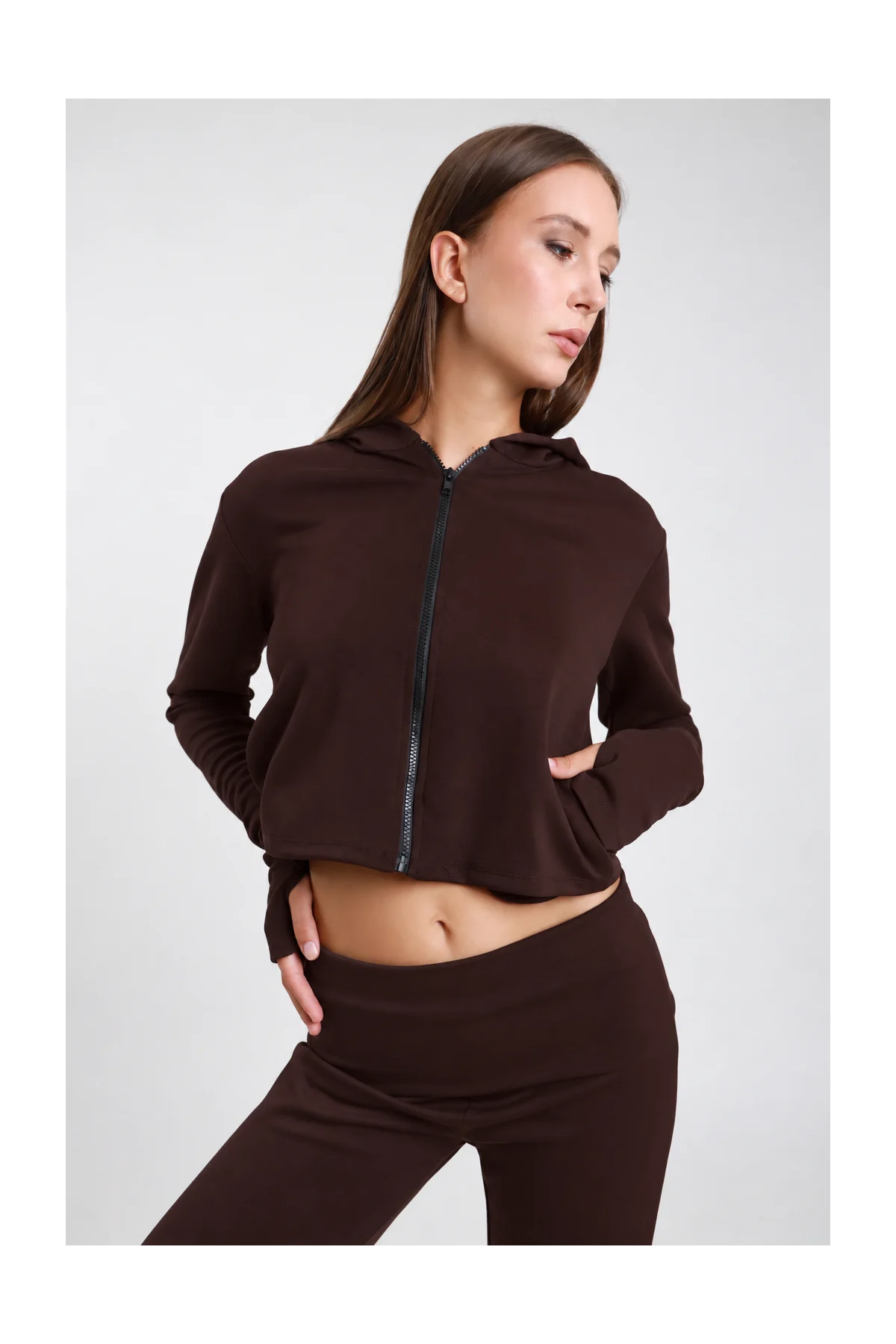 Felpa Crop Zip DAYTHRU marrone cioccolato scuro cappuccio zip nera frontale logo SMX manica orlo crop polsino foro pollice crop fit busto frontale 3/4 mano tasca sguardo basso - modella busto frontale 3/4 mano tasca sguardo basso capelli castani lisci sfondo bianco coordinato marrone scuro pantalone wide-leg