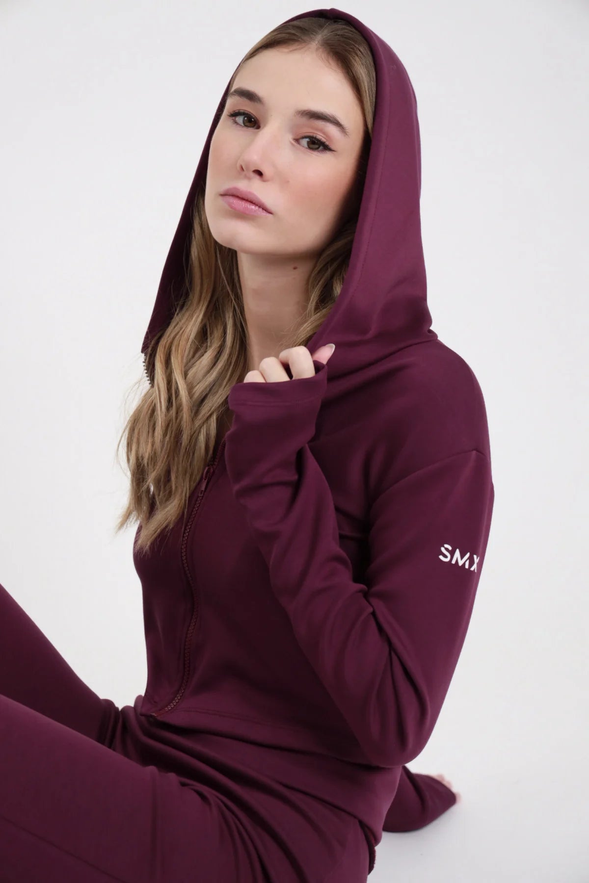 Felpa Crop Zip SMX Limited Edition bordeaux cappuccio ampio coulisse logo SMX bianco braccio sinistro ben visibile thumbhole polsino zip frontale seduta 3/4 mano cappuccio - modella seduta 3/4 mano cappuccio sguardo morbido capelli mossi castani sfondo bianco pantaloni flare SMX bordeaux coordinati abbinati