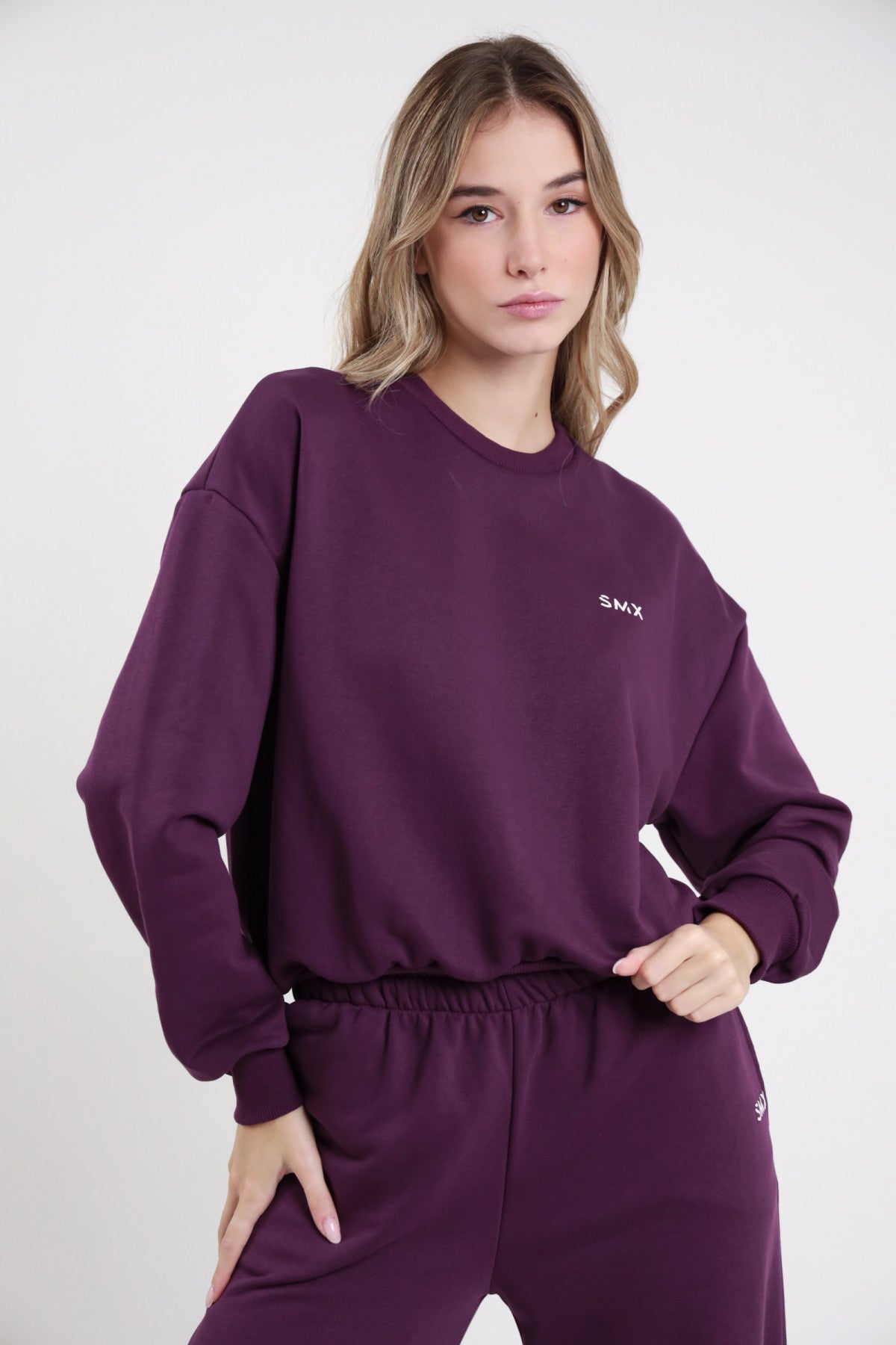 Felpa Girocollo SMX Limited Edition viola melanzana 65% cotone french terry girocollo crewneck logo SMX bianco petto sinistro manica balloon oversize polsino costina bordo costina boxy frontale busto mani fianchi - modella busto frontale mani fianchi sguardo frontale capelli biondi mossi sfondo bianco pantalone wide leg viola coordinato abbinato