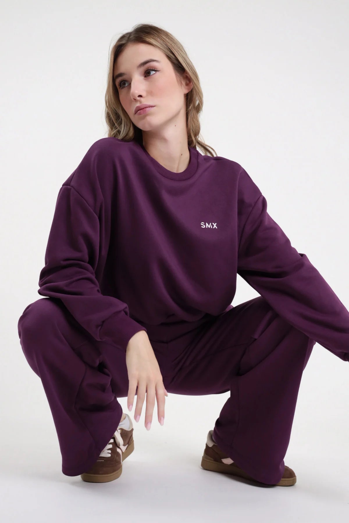 Felpa Girocollo SMX Limited Edition viola melanzana logo SMX petto crewneck manica balloon oversize bordo costina accovacciata frontale 3/4 laterale mano gamba - modella accovacciata frontale 3/4 laterale mano gamba sguardo laterale capelli biondi mossi sfondo bianco pantalone wide leg viola coordinato sneakers marrone tan abbinati