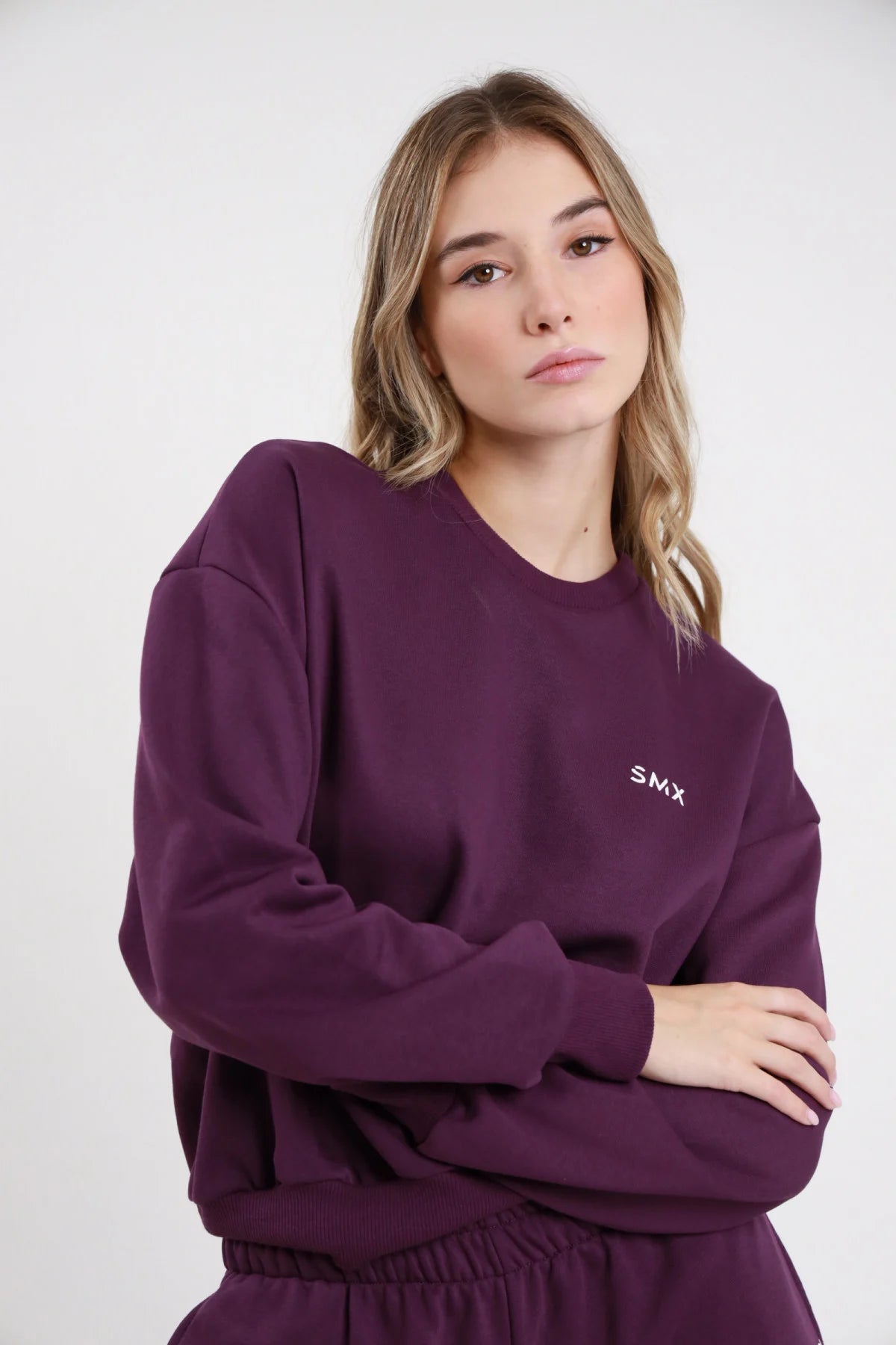 Felpa Girocollo SMX Limited Edition viola melanzana logo SMX petto sinistro ricamato bianco girocollo crewneck manica balloon bordo costina frontale busto braccia conserte - modella busto frontale braccia conserte sguardo frontale capelli biondi mossi sfondo bianco pantalone wide leg viola coordinato abbinato