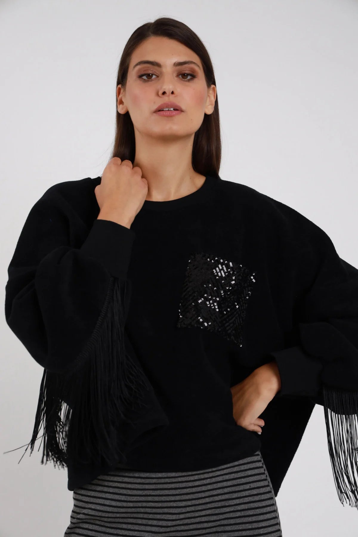 Felpa Oversize Frange Tasca Paillettes nera girocollo frange lunghe nere maniche retro tasca paillettes fiore nero tono su tono silhouette boxy - modella busto frontale mano al collo sguardo intenso frontale capelli castani lisci sfondo bianco gonna midi righe grigia coordinata