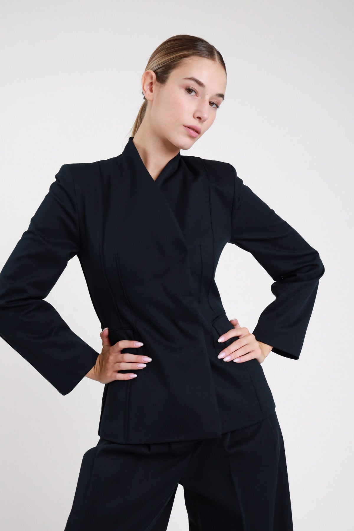 Blazer asimmetrico nero nervature verticali - modella busto frontale mani sui fianchi con pantaloni neri coordinati
