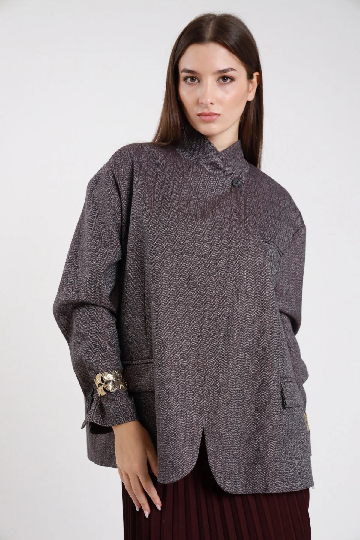 Blazer spigato herringbone grigio antracite marrone scuro lurex bronzo ramato frontale intera collo alto funnel neck abbottonatura asimmetrica singolo bottone scuro manica balloon bishop bracciale gioiello floreale foglia oro polso tasche laterali patch corpo oversize boxy orlo asimmetrico curved - modella frontale intera sguardo frontale capelli castani lisci sfondo bianco gonna plissé bordeaux
