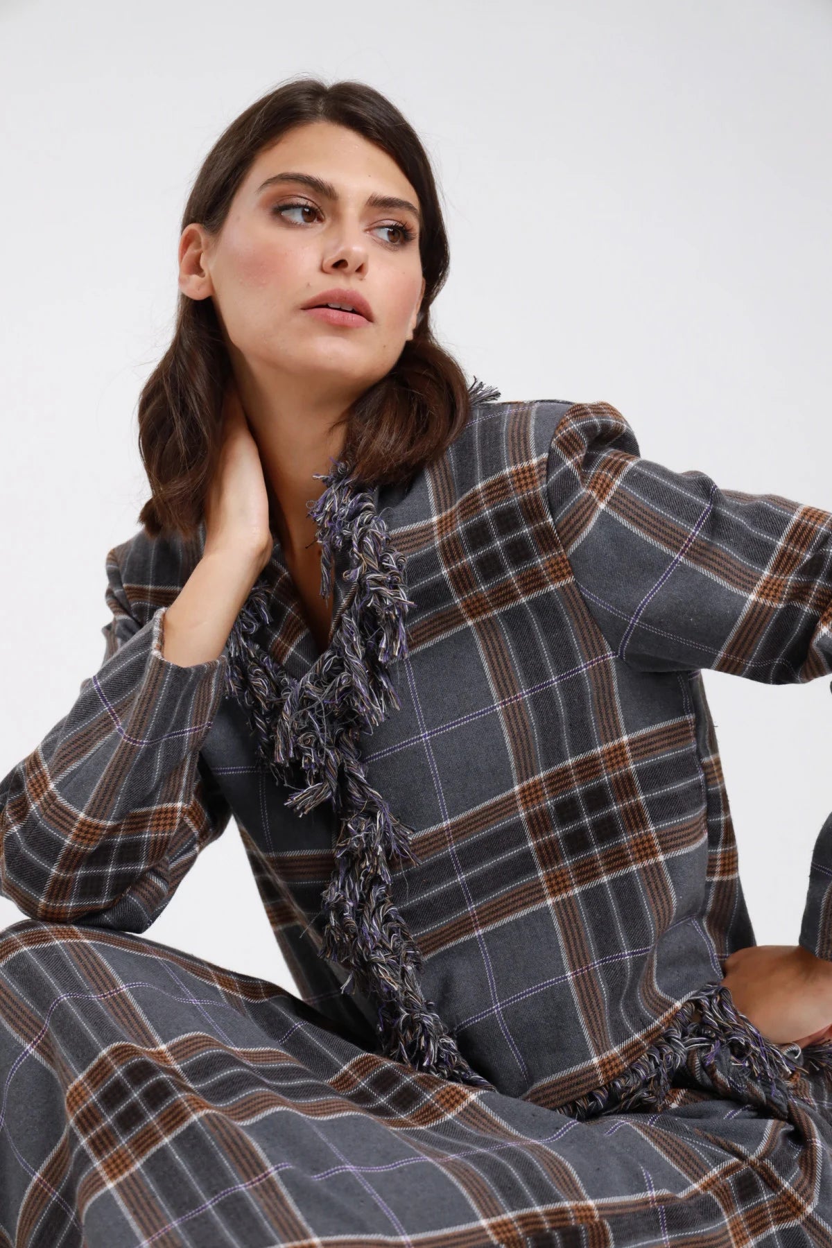 Giacca check tartan grigio antracite marrone donna boxy cropped frange multifilo bordo frontale V fondo spalle strutturate - dettaglio frontale mezzo busto seduta mano al collo con gonna check coordinata