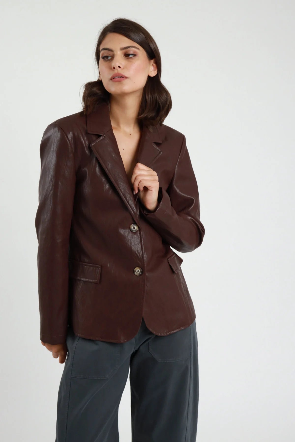 Blazer Monopetto Ecopelle marrone cioccolato texture crinkled vintage revers V notched 2 bottoni tasche patta spalle strutturate busto frontale mano revers - modella busto frontale mano revers sguardo laterale capelli castani sfondo bianco pantaloni grigio antracite
