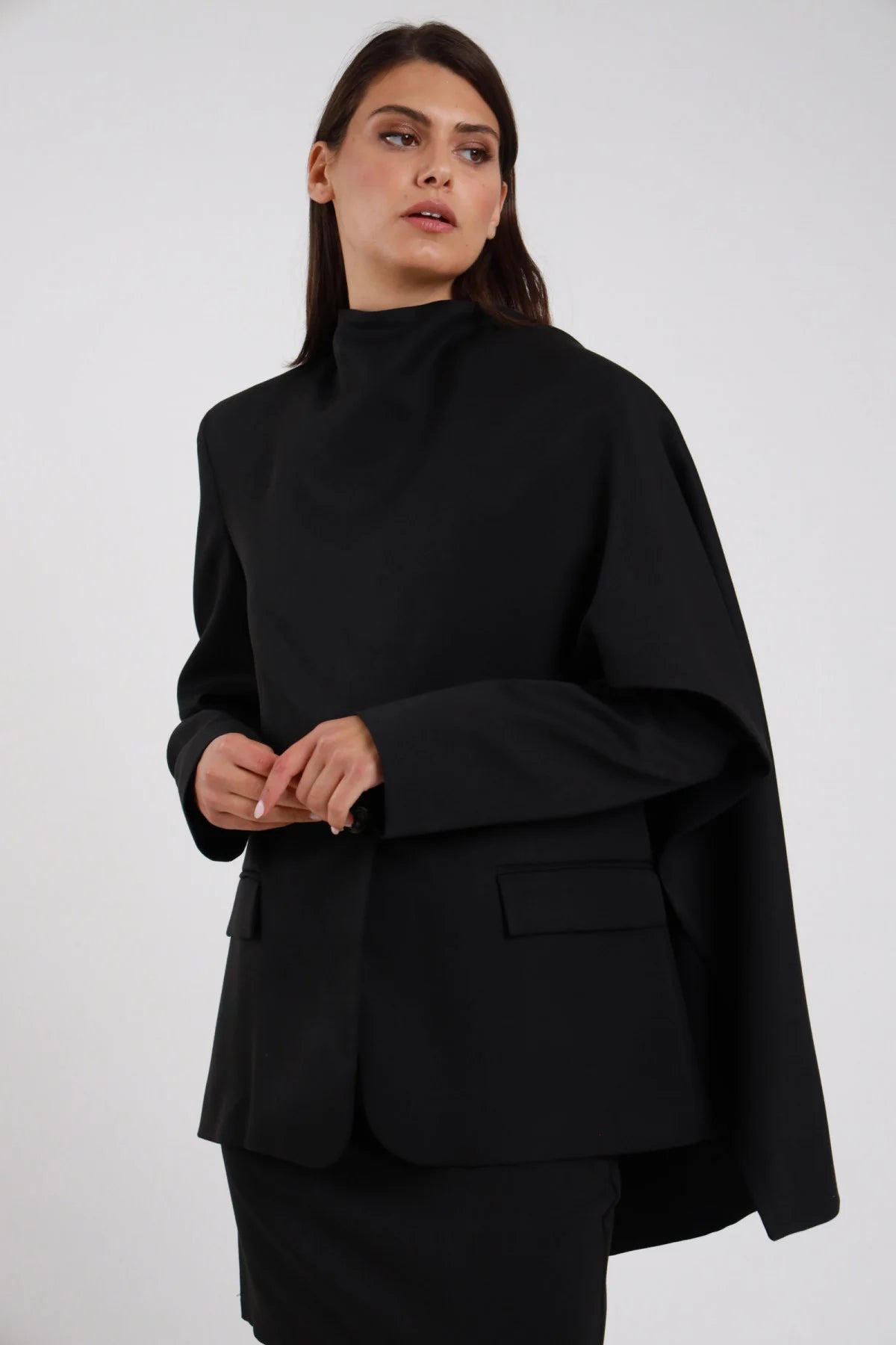 Blazer Scialle nero colletto alto scialle mantella drappeggiata laterale nera monobottone tasca pattina spalle strutturate busto frontale 3/4 mani intrecciate sguardo laterale - modella busto frontale 3/4 mani intrecciate sguardo laterale capelli castani lisci sfondo bianco minigonna nera coordinata
