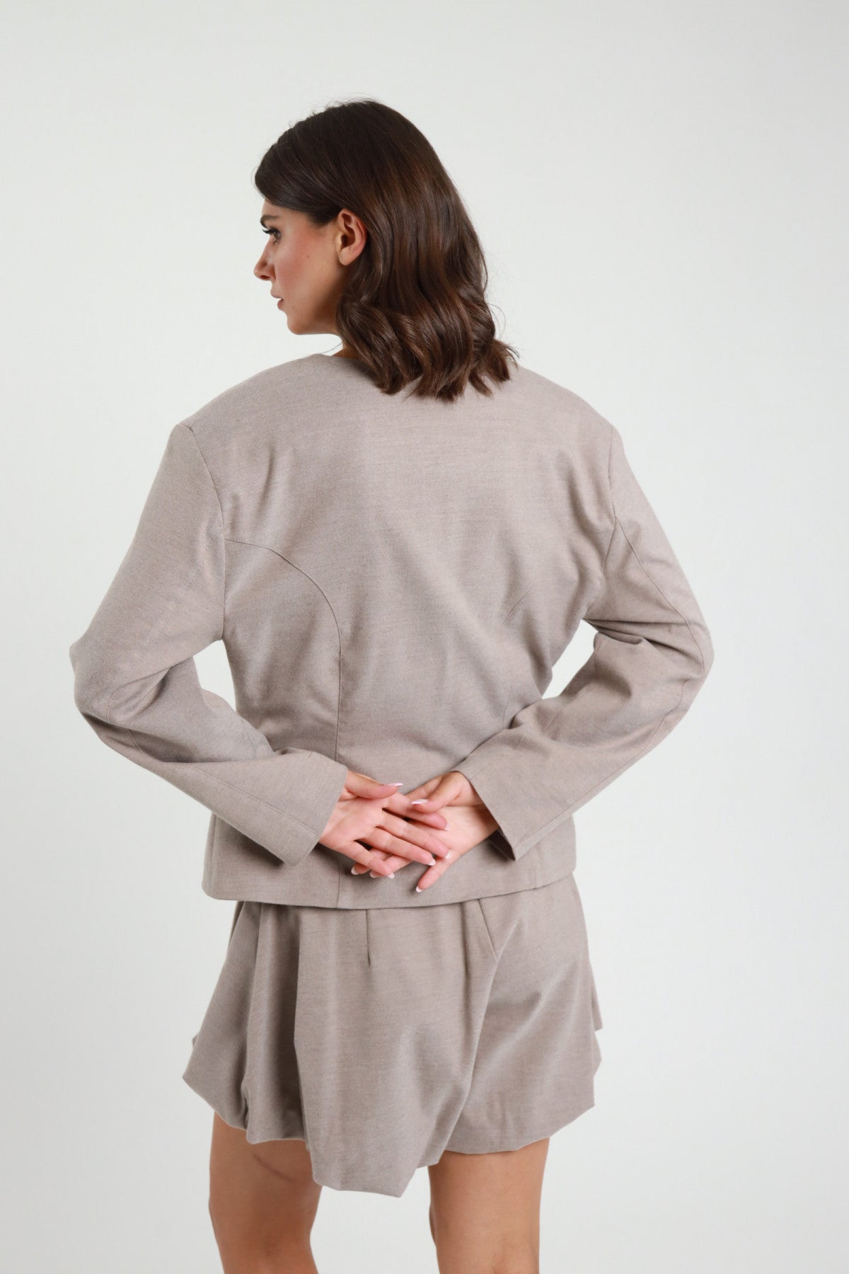 Giacchina crop sartoriale tortora greige collo coreana Mao bottoni monopetto ton sur ton cuciture vista retro busto mani giunte schiena - modella retro busto mani giunte schiena sguardo laterale capelli castani sfondo bianco short coordinato tortora