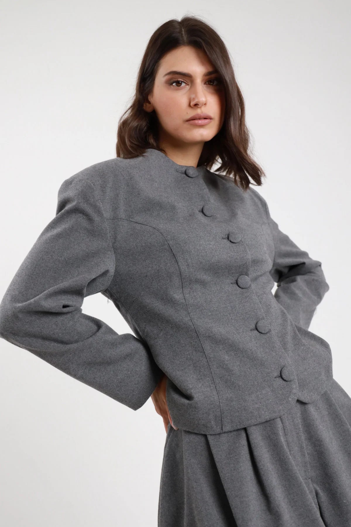 Giacchina crop sartoriale grigio melange antracite collo coreana Mao bottoni doppiopetto ton sur ton cuciture vista spalle strutturate busto frontale 3/4 mani fianchi - modella busto frontale 3/4 mani fianchi sguardo frontale capelli castani sfondo bianco short coordinato grigio stivali neri