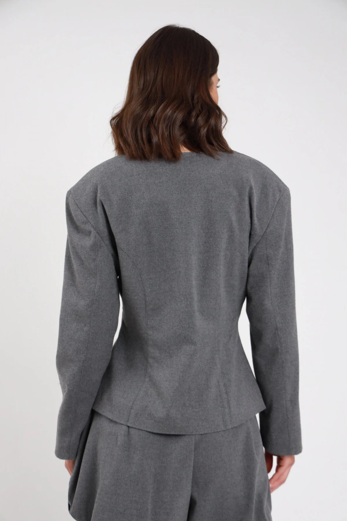 Giacchina crop sartoriale grigio melange antracite collo coreana Mao bottoni doppiopetto ton sur ton cuciture vista retro busto - modella retro busto mani fianchi sguardo laterale capelli castani sfondo bianco short coordinato grigio stivali neri