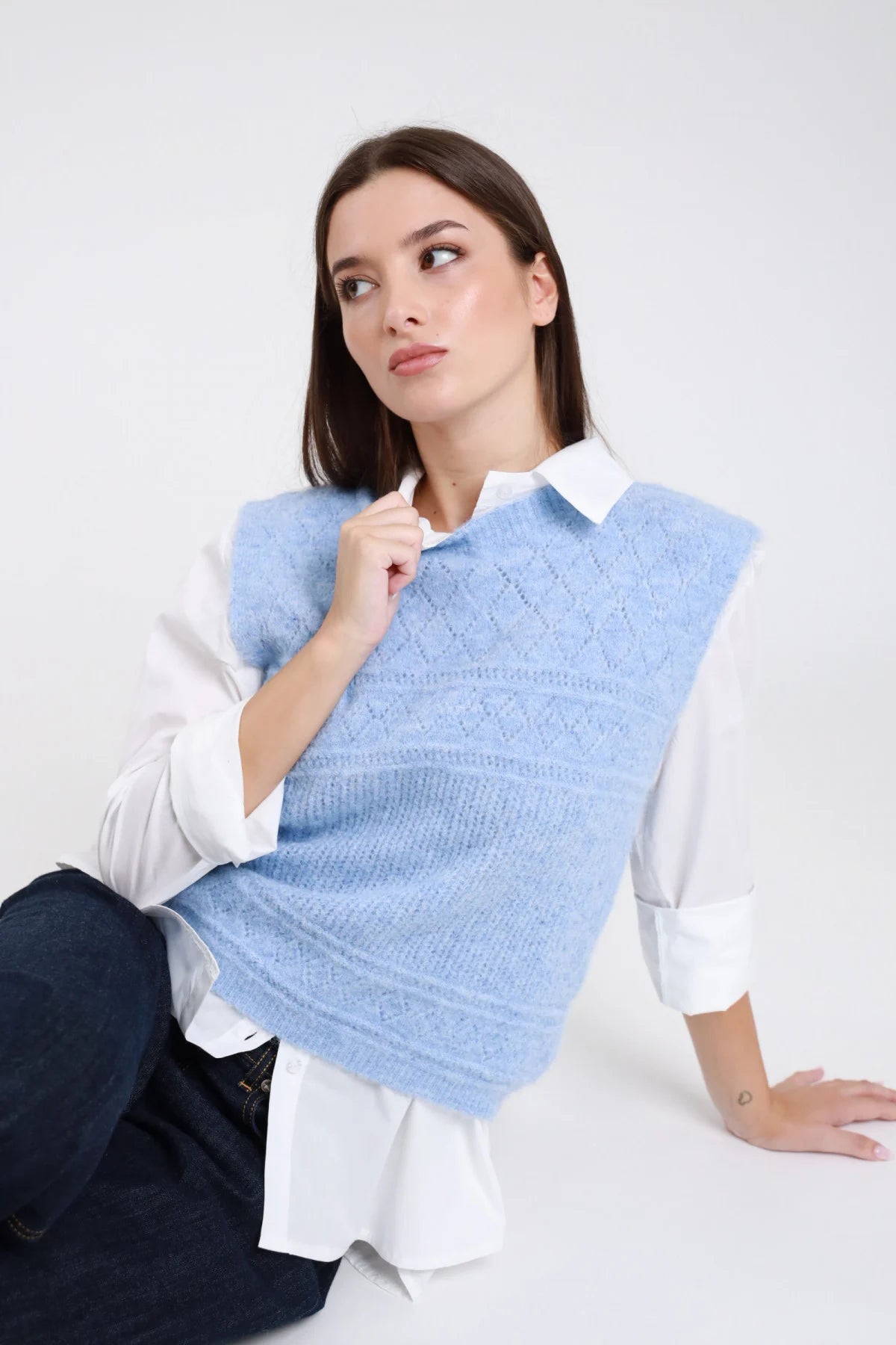 Gilet maglia eyelash bouclé traforato rombi senza maniche girocollo crop azzurro celeste baby blue frontale seduta mano scollo camicia bianca jeans blu - modella frontale seduta mano scollo sguardo laterale capelli castani scuri lisci sfondo bianco camicia bianca jeans blu scuro