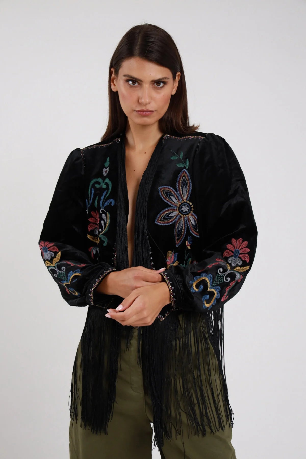 Giacchino Ricamato Frange nero velluto raso ricamo floreale folk multicolor fiore centrale petali volute colorate frange lunghe nere bordi dorati dettaglio frontale busto frontale 3/4 mani giunte sguardo camera - modella busto frontale 3/4 mani giunte sguardo camera capelli castani scuri sfondo bianco pantaloni verde militare