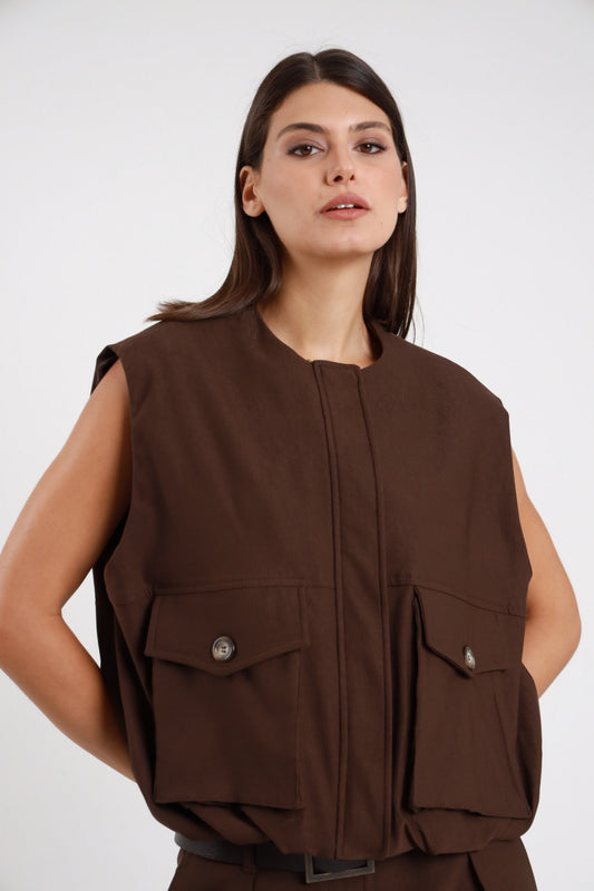 Gilet Cargo Tasconi marrone cioccolato 78% viscosa collo coreana basso zip frontale nascosta patta 2 tasche cargo petto bottone brunito spalle strutturate silhouette oversize boxy frontale busto - modella busto frontale mani tasche fianchi sguardo laterale alto make-up bold capelli castani lisci sfondo bianco pantalone tasconi marrone coordinato cintura ecopelle nera