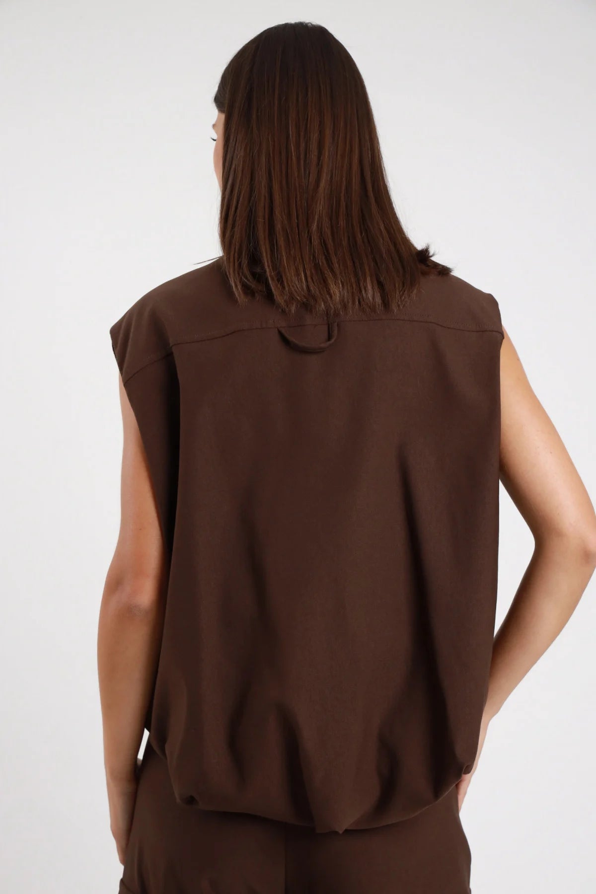 Gilet Cargo Tasconi marrone cioccolato 78% viscosa retro liscio cuciture verticali sartoriali silhouette oversize boxy posteriore intera - modella intera posteriore posa mani fianchi sguardo basso capelli castani lisci sfondo bianco pantalone tasconi marrone coordinato
