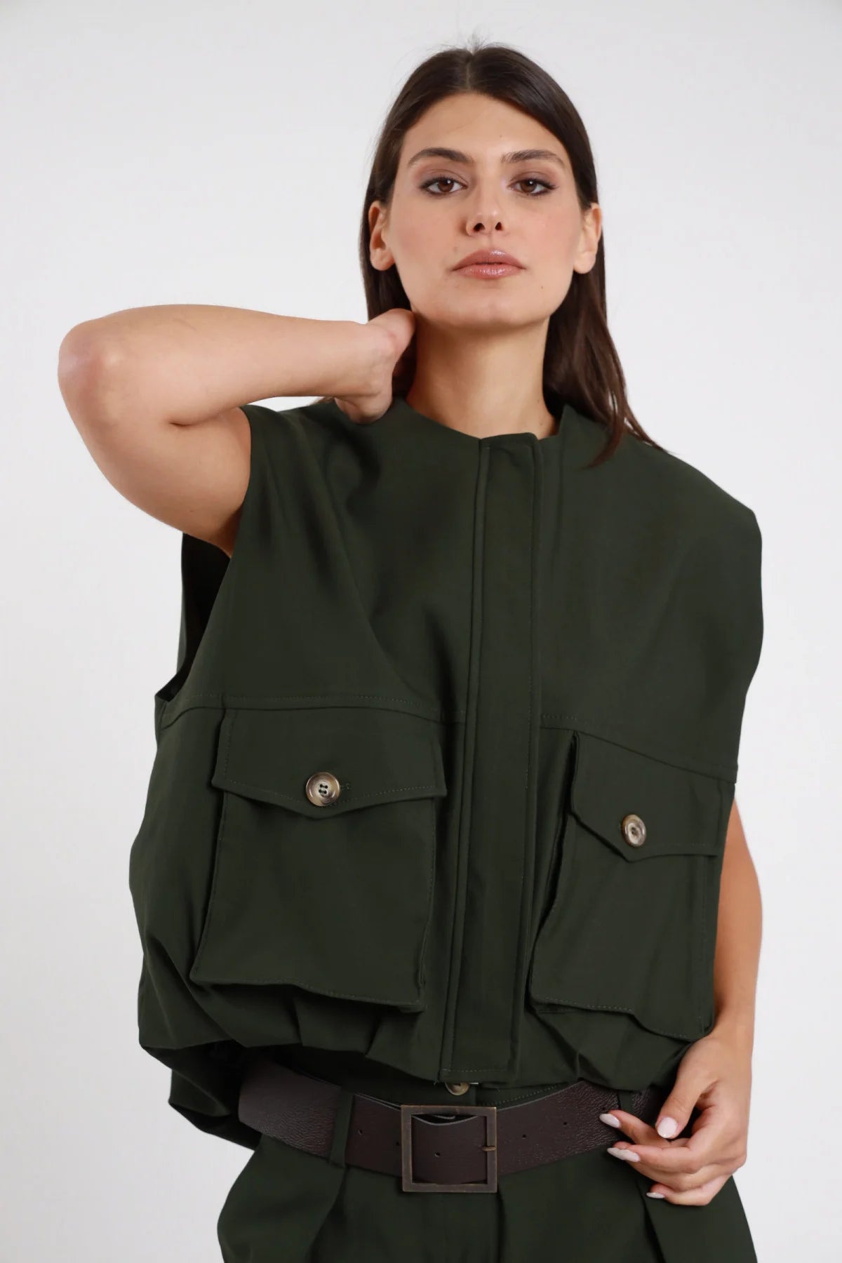 Gilet Cargo Tasconi verde militare bottiglia 78% viscosa collo coreana zip nascosta patta 2 tasche cargo petto bottone brunito spalle strutturate silhouette oversize boxy frontale busto - modella busto frontale posa mano collo sguardo frontale intenso capelli castani lisci sfondo bianco pantalone tasconi verde militare coordinato cintura ecopelle marrone
