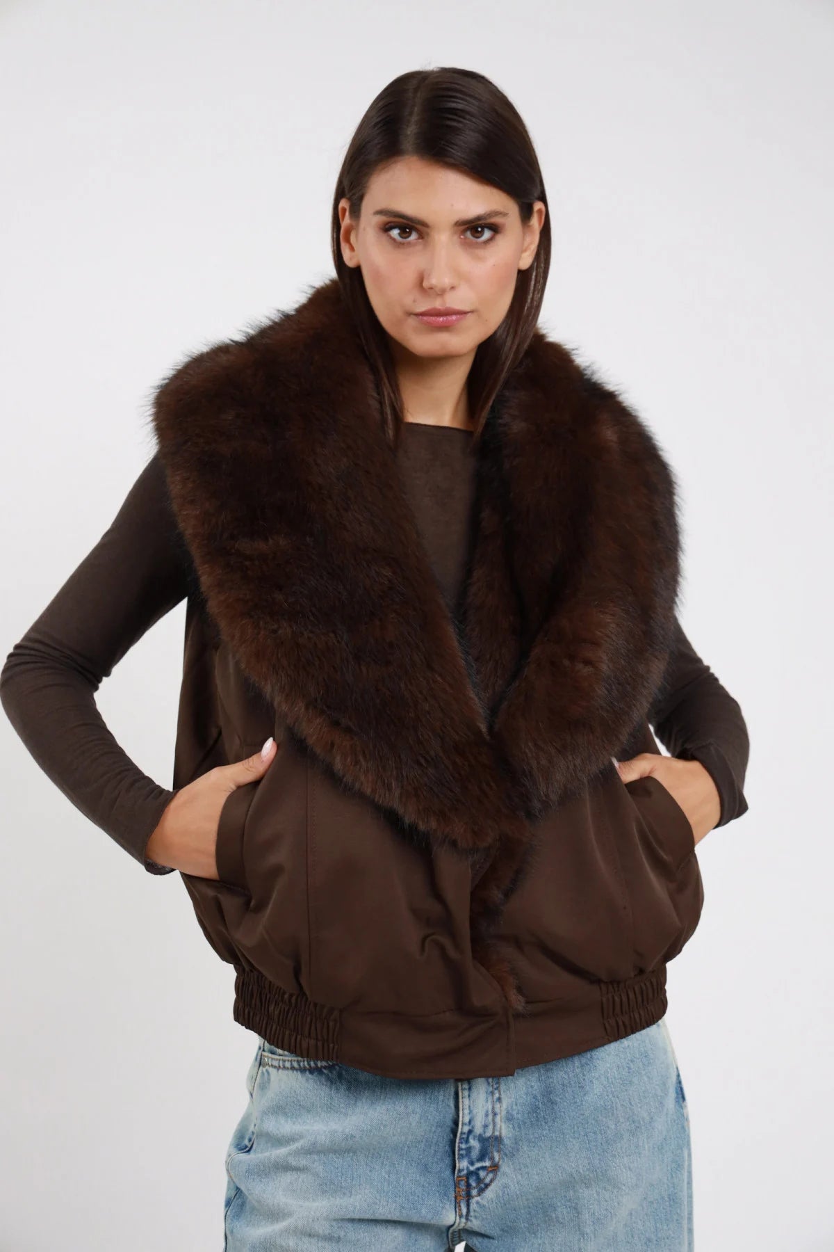 Gilet Ecopelliccia marrone cioccolato scuro colletto maxi ecopelliccia corpo scamosciato suede elastico vita tasche frontali busto frontale 3/4 mani tasche - modella busto frontale 3/4 mani tasche sguardo camera capelli castani lisci sfondo bianco maglia marrone manica lunga jeans chiari
