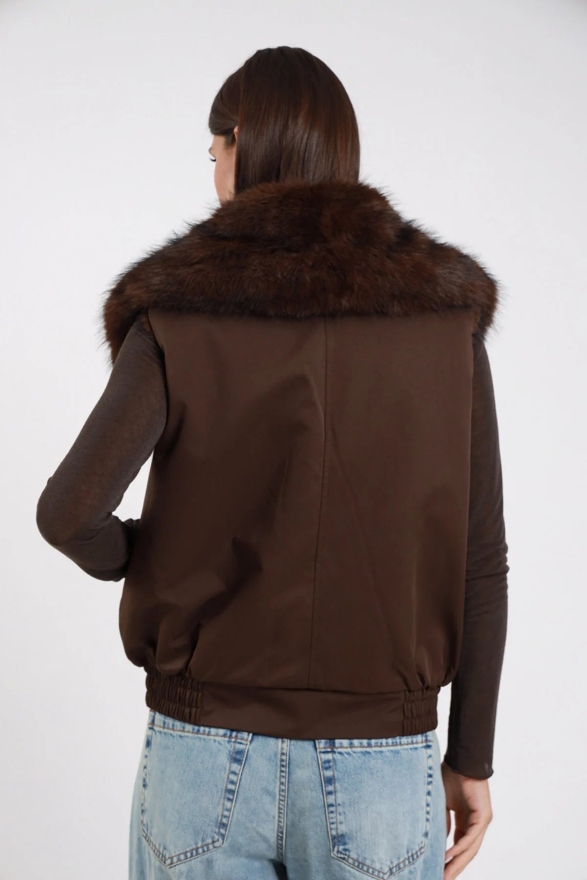 Gilet Ecopelliccia marrone cioccolato scuro colletto maxi ecopelliccia corpo scamosciato suede retro intera schiena collare voluminoso - modella retro intera capelli castani lisci sfondo bianco maglia marrone manica lunga jeans chiari