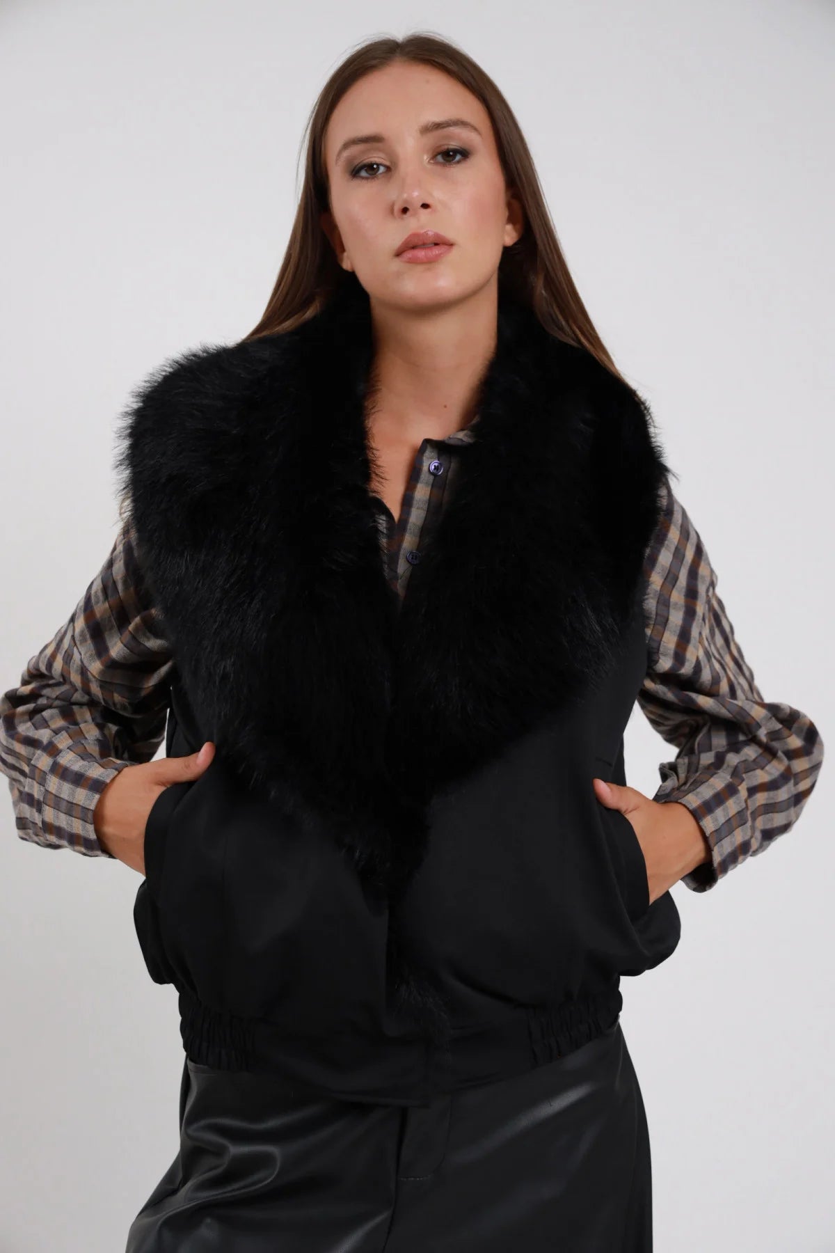Gilet Ecopelliccia nero colletto maxi ecopelliccia nera corpo scamosciato suede nero monobottone busto frontale 3/4 mani tasche sguardo camera - modella busto frontale 3/4 mani tasche sguardo camera capelli castani lisci sfondo bianco camicia scozzese beige grigia gonna nera ecopelle
