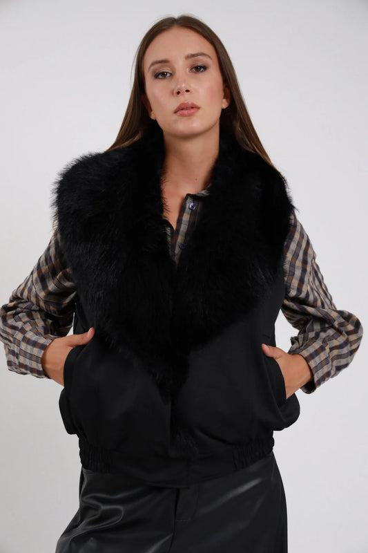 Gilet Ecopelliccia nero colletto maxi ecopelliccia nera corpo scamosciato suede nero monobottone busto frontale 3/4 mani tasche sguardo camera - modella busto frontale 3/4 mani tasche sguardo camera capelli castani lisci sfondo bianco camicia scozzese beige grigia gonna nera ecopelle