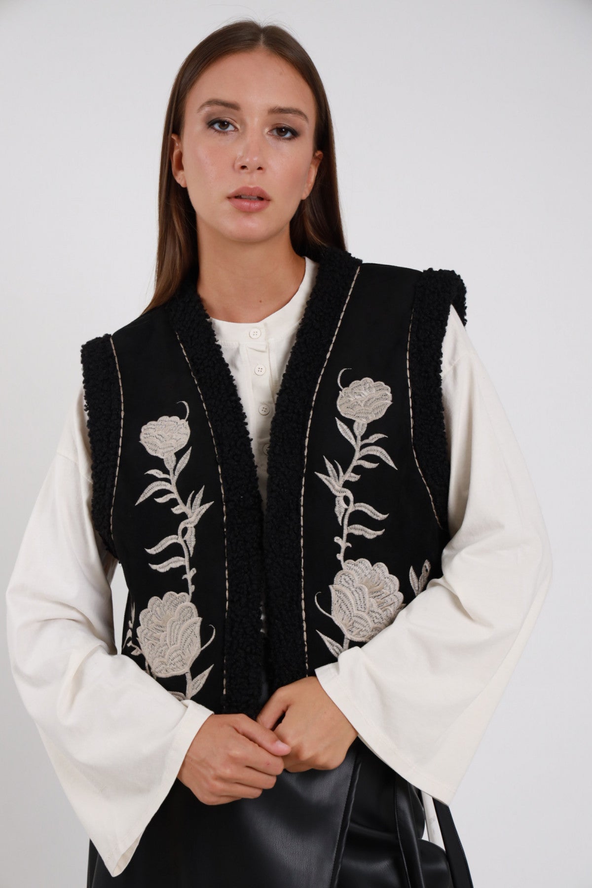 Gilet Ecomontone Ricamato nero ricamo floreale rose foglie avorio grigio perla bordi bouclé ricci scollo V aperto silhouette crop busto frontale 3/4 mani giunte sguardo camera - modella busto frontale 3/4 mani giunte sguardo camera capelli castani lisci sfondo bianco camicia bianca manica ampia gonna ecopelle nera