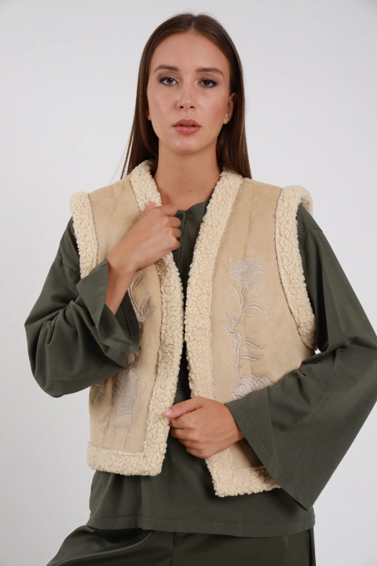 Gilet Ecomontone Ricamato panna avorio lato sherpa ecomontone caldo voluminoso bordi sherpa bouclé tono su tono ricamo floreale panna suede scollo V aperto busto frontale 3/4 mano colletto sguardo camera - modella busto frontale 3/4 mano colletto sguardo camera capelli castani lisci sfondo bianco felpa verde oliva pantaloni verde oliva